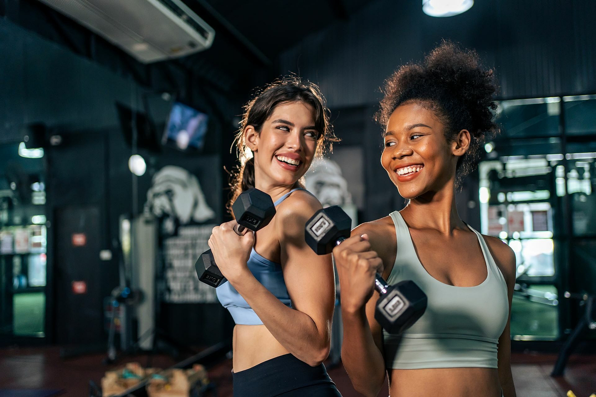 Two women enthusiastically lifting dumbbells in a vibrant gym setting, exuding energy and positivity.