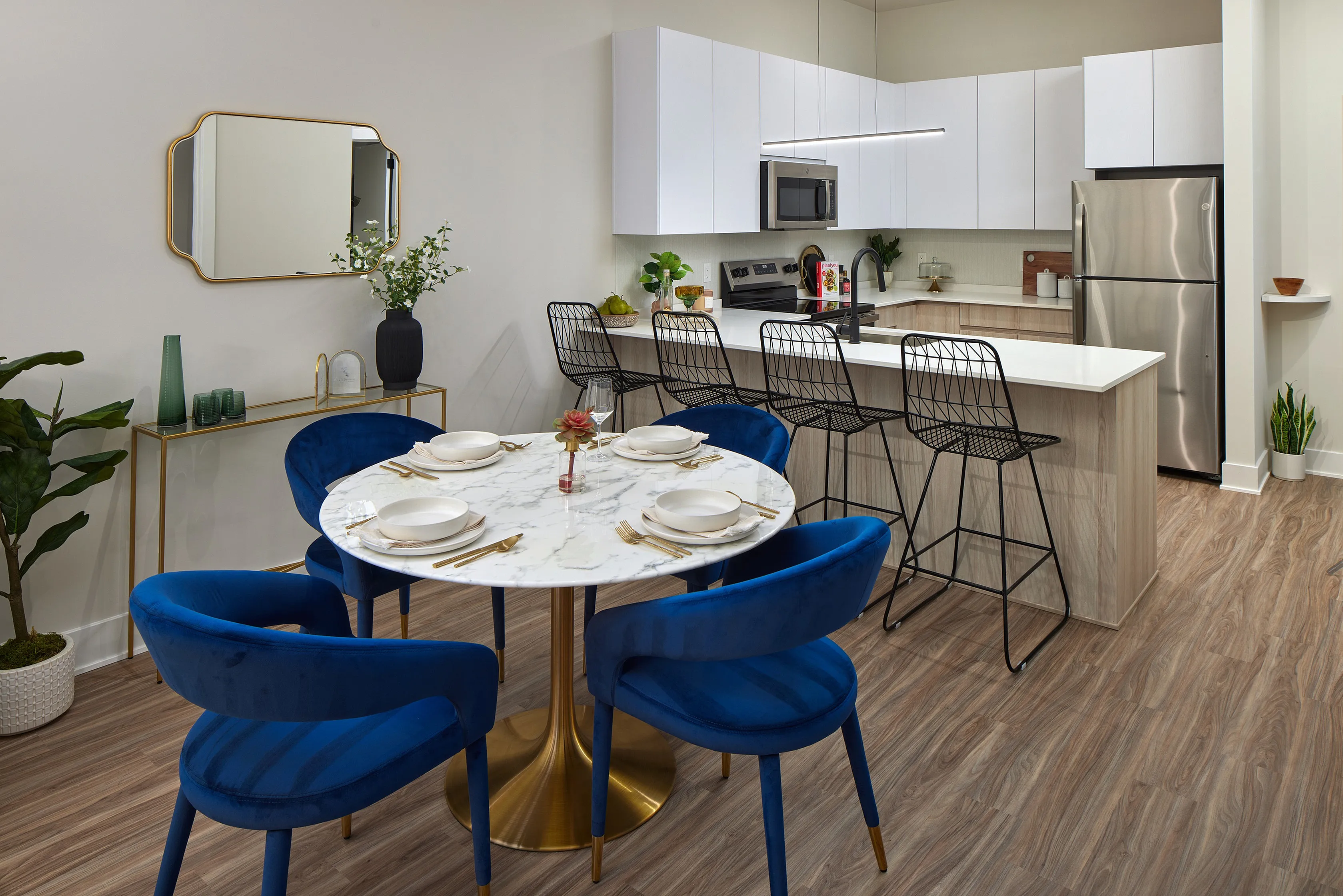 Elegant modern kitchen and dining area featuring a marble dining table with chic blue velvet chairs, sleek white cabinetry, and a stylish breakfast bar with high chairs.