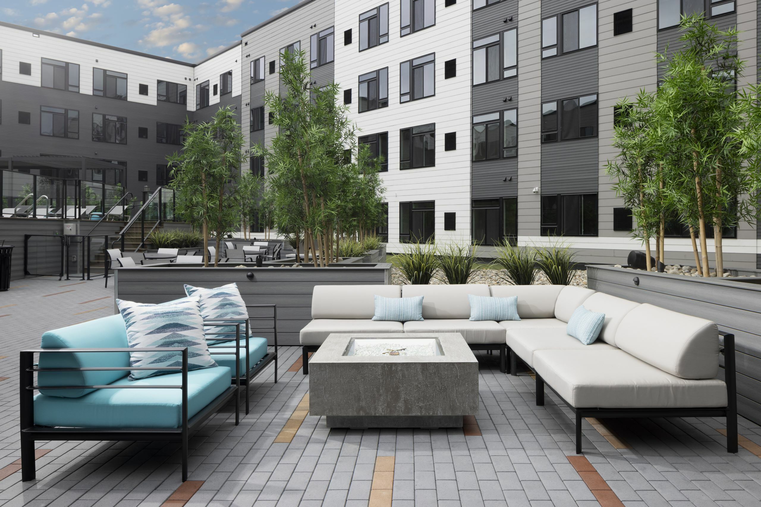 Modern apartment complex courtyard featuring stylish outdoor seating, lush greenery, and a sleek fire pit under a picturesque sky.