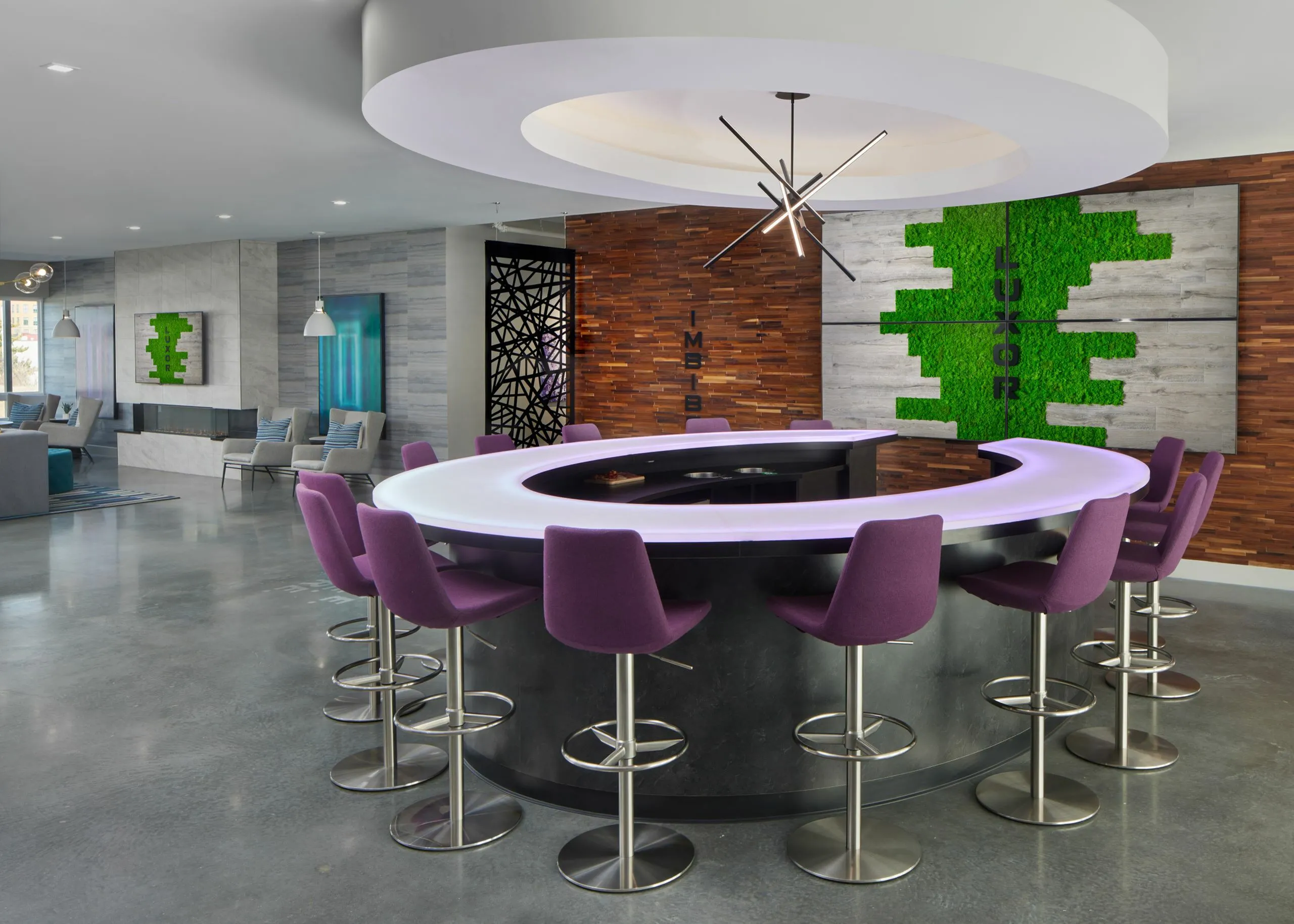Modern communal lounge featuring a chic circular bar with vibrant purple chairs, accentuated by stylish lighting and lush green wall art.