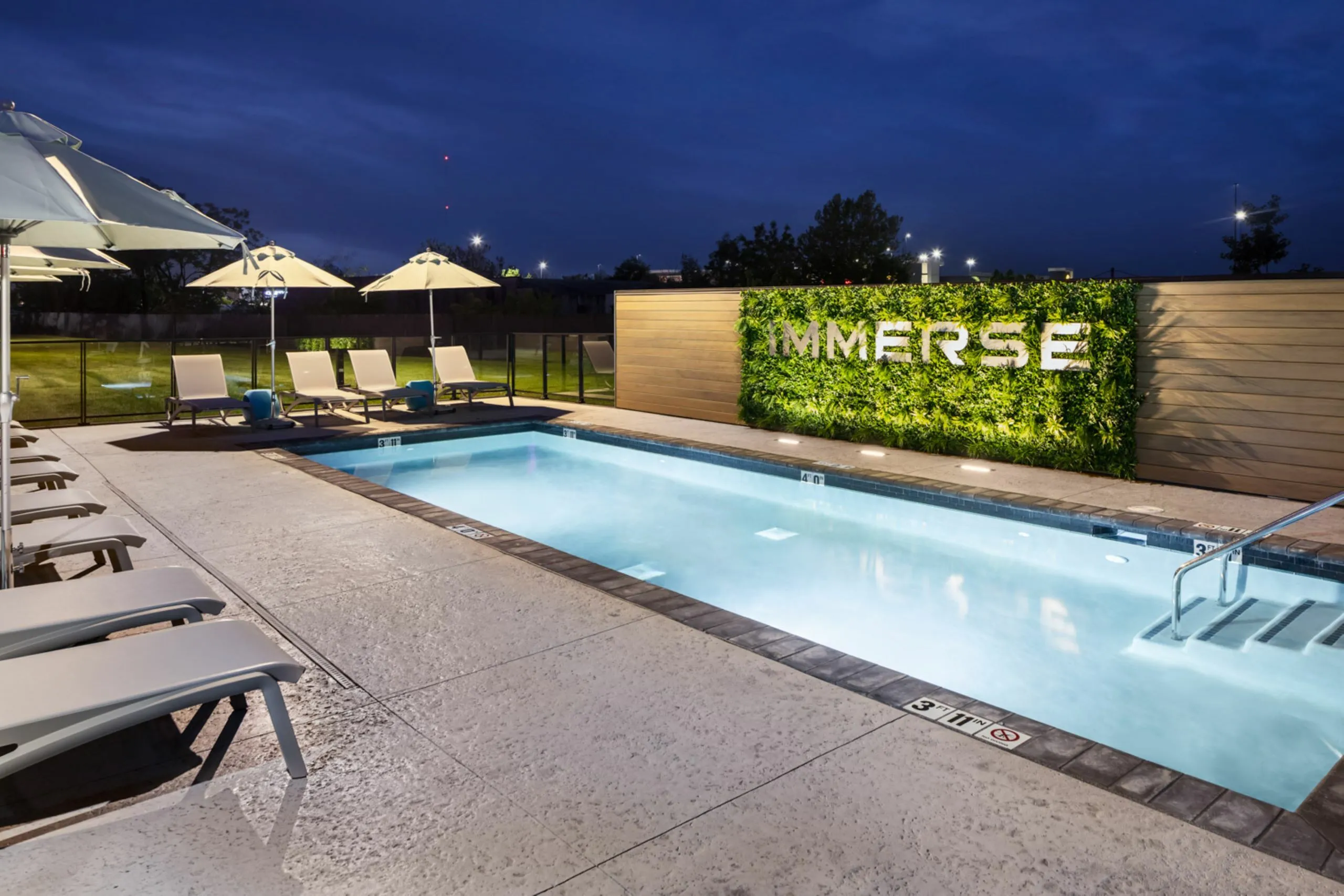 Luxurious night view of an illuminated swimming pool surrounded by elegant lounge chairs and umbrellas, set against a lush green wall.