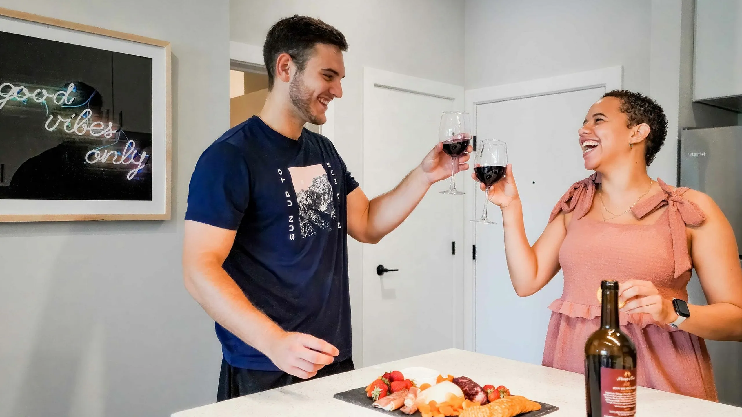 Happy couple toasting with wine in a modern kitchen featuring a charcuterie board and a 'good vibes only' neon sign.