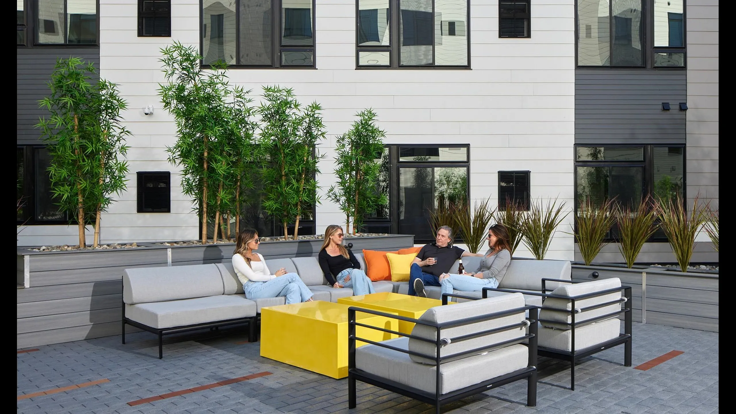 Chic outdoor lounge area with modern seating and vibrant yellow table, surrounded by lush greenery and contemporary apartment architecture.