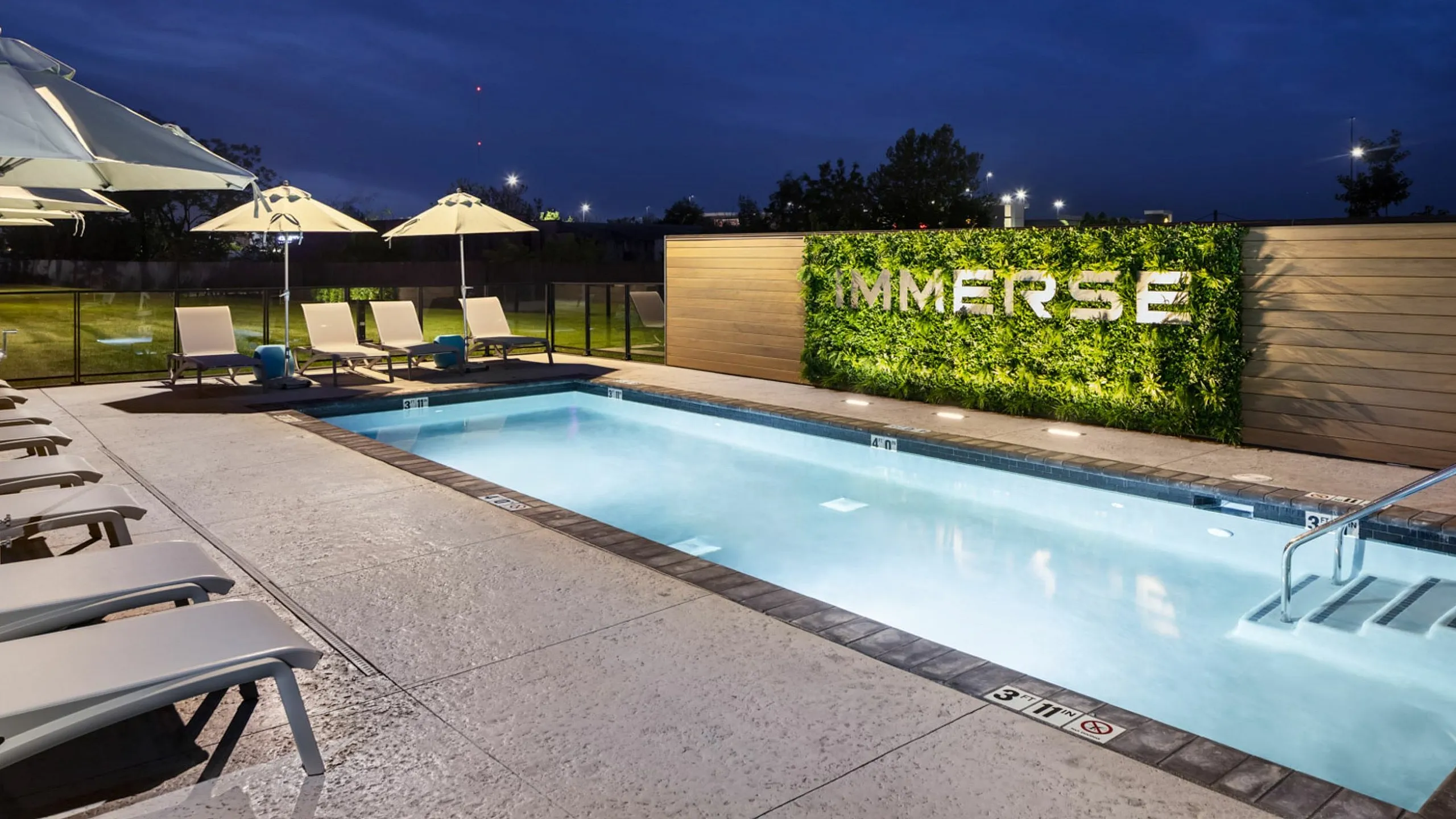Elegant pool area at night with ambient lighting, stylish lounge chairs, and lush green wall at 'Immerse'.