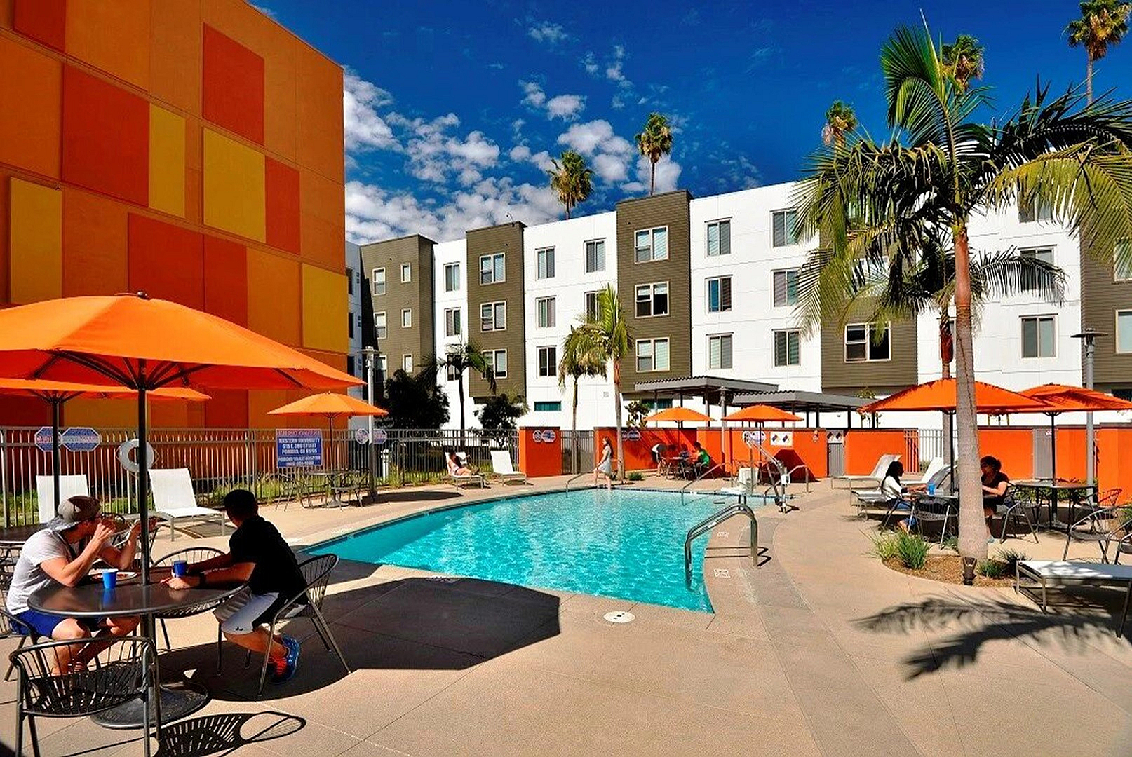 Vibrant apartment complex with a luxurious pool area, surrounded by tropical palm trees and bright orange accents, offering an enticing outdoor oasis for relaxation and leisure.