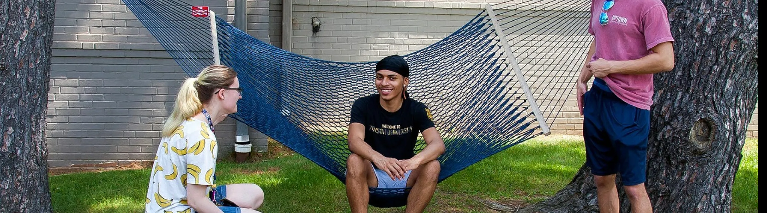 Three friends enjoying a conversation in a hammock setup between trees in a sunny yard.