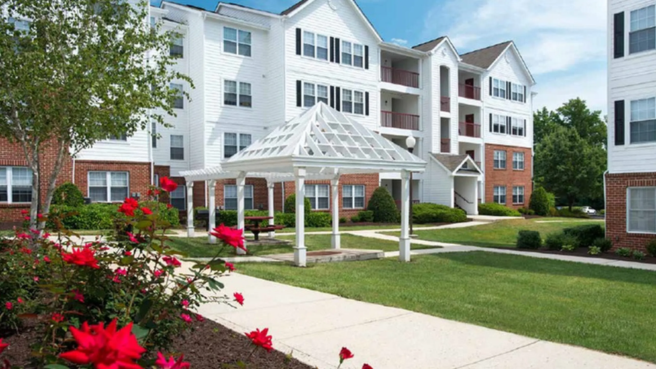 Beautiful multi-story apartment building with white facade and red brick accents surrounded by lush greenery and vibrant flowers.