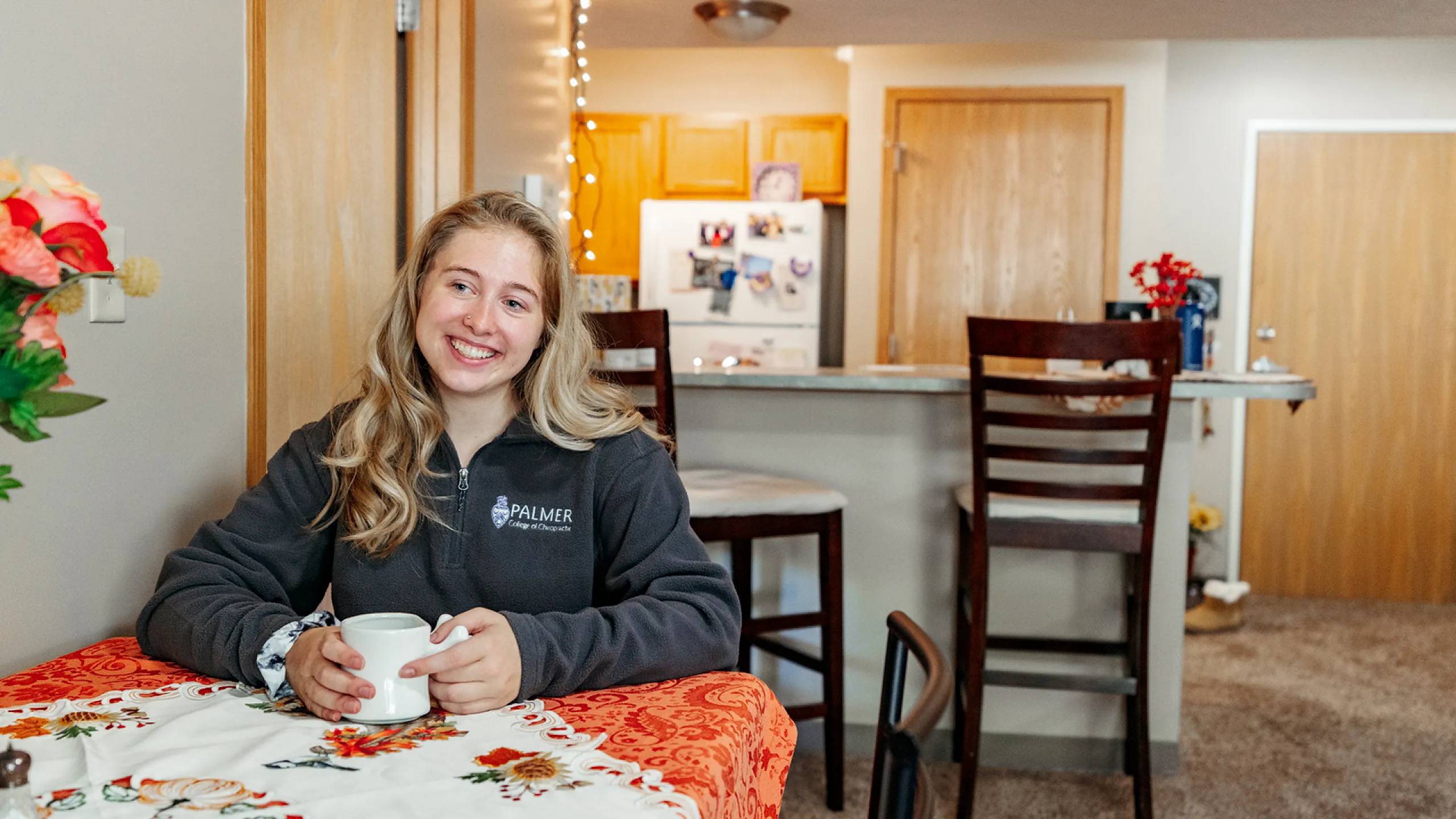 Woman enjoying a cozy kitchen area with a warm drink, featuring a table with floral decor and a view of a modern kitchen.