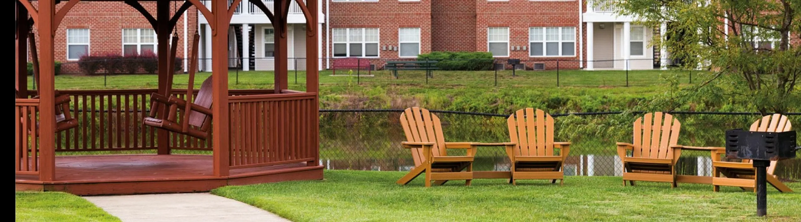 Charming outdoor space with a wooden gazebo and Adirondack chairs overlooking a serene pond near red-brick apartments.