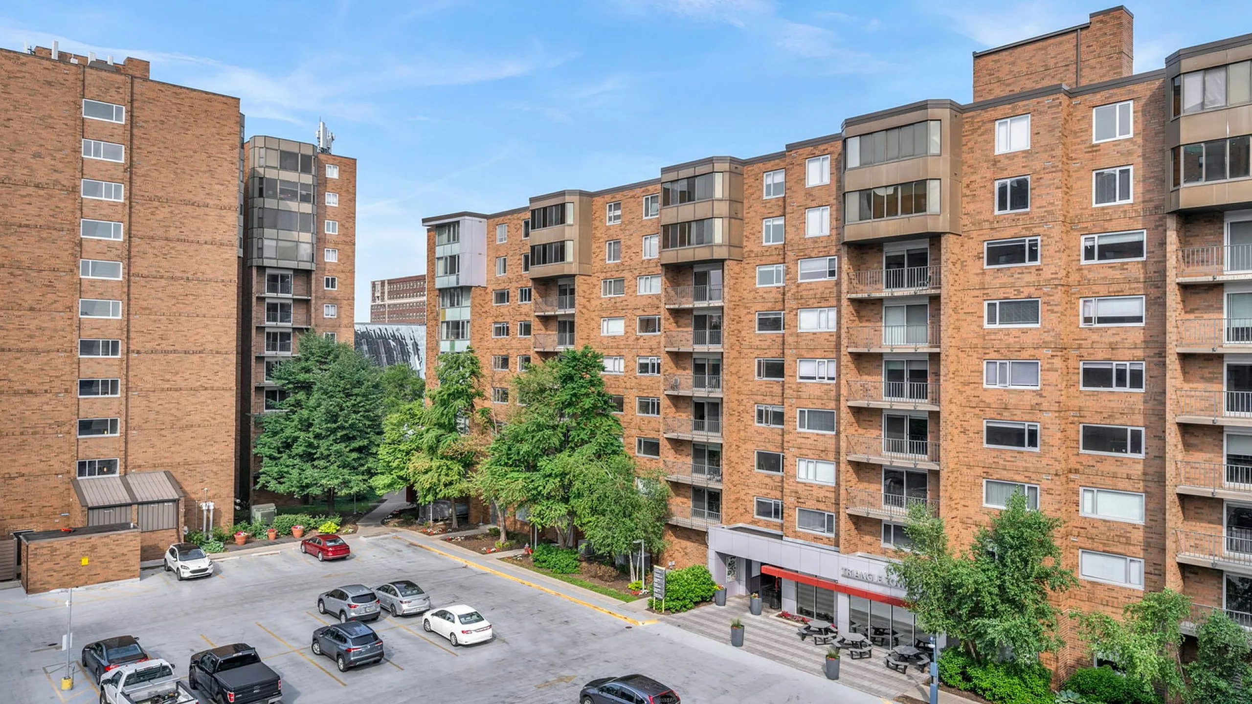 Urban residential buildings with parking lot and lush green trees under a clear blue sky.