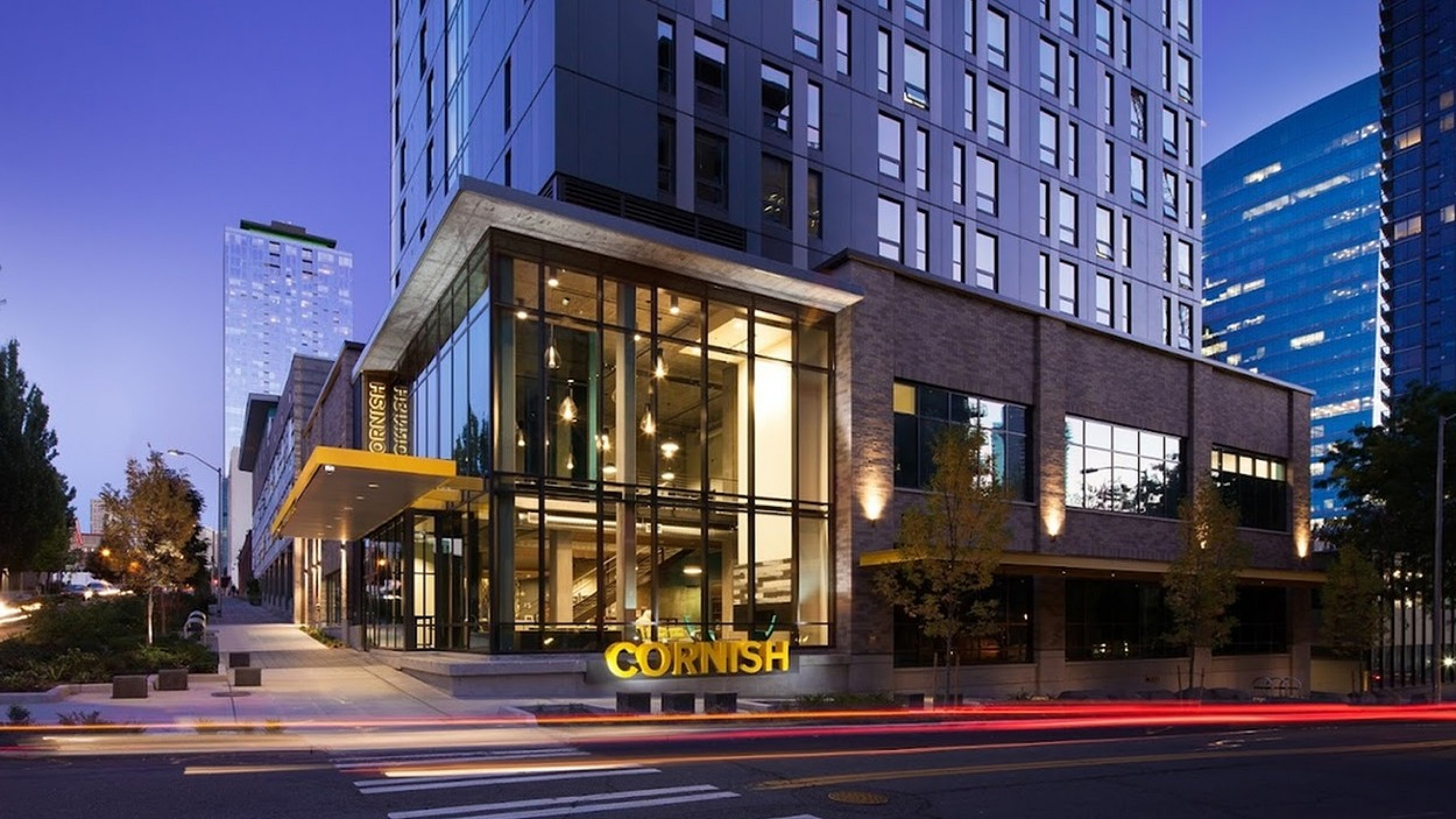 Stunning modern high-rise building with sleek glass facade illuminated against a twilight sky, featuring a welcoming, brightly-lit entrance.