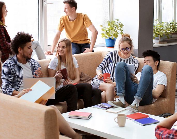 Vibrant communal lounge with diverse group of young adults relaxing and socializing on cozy sofas, enjoying a bright, plant-filled environment.