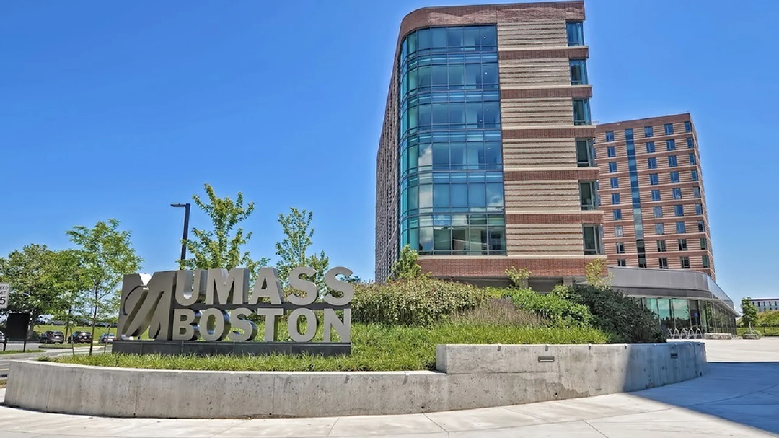 Modern university campus with striking architecture, featuring expansive glass windows and a prominent UMass Boston sign under a clear blue sky.