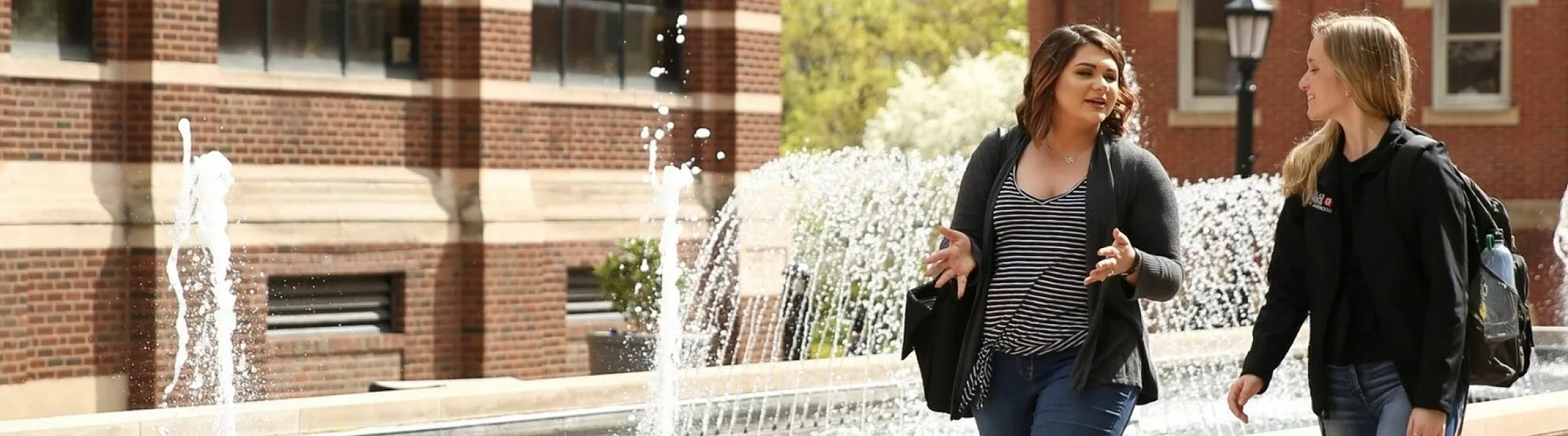 Two smiling women enjoying a sunny day walking by a charming brick fountain in an urban setting.
