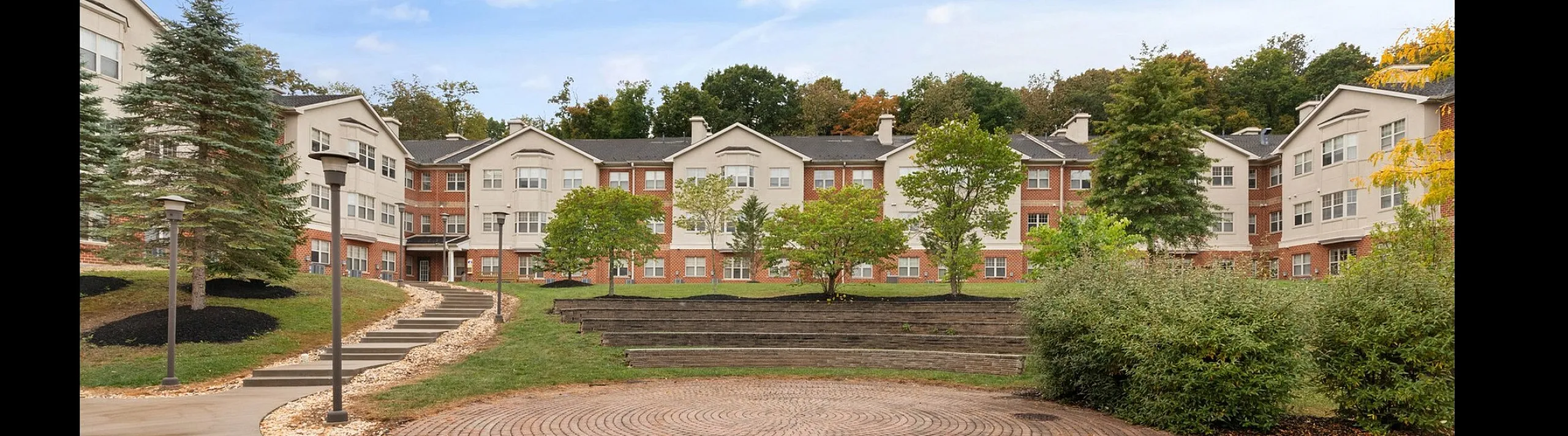 Charming apartment complex with a beautifully landscaped entrance, featuring lush greenery, a circular brick patio, and modern architecture under a bright blue sky.
