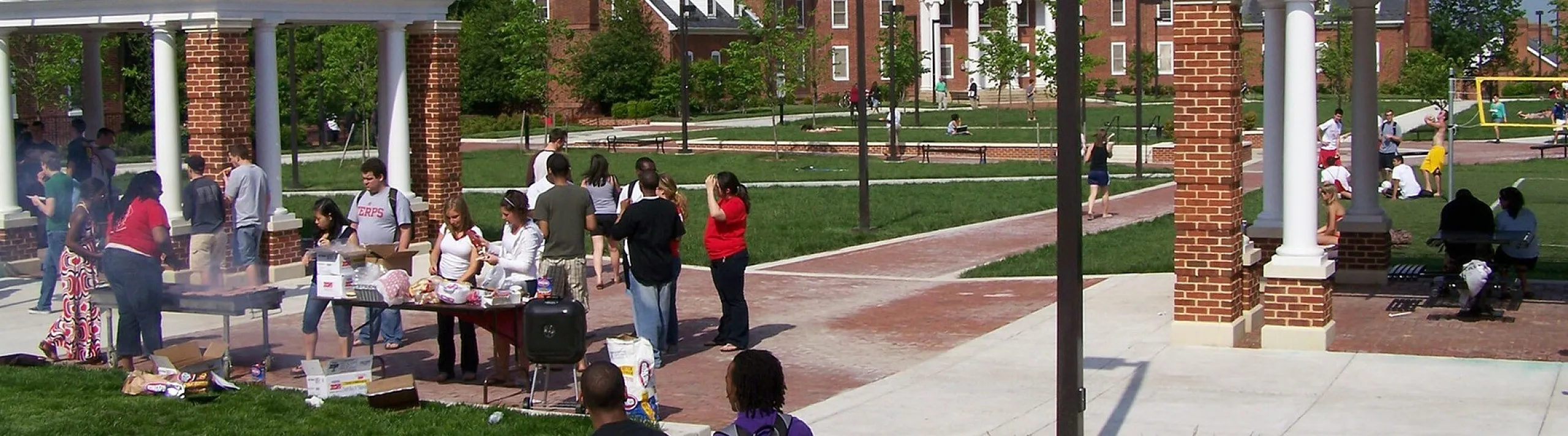Vibrant community gathering in a scenic outdoor campus courtyard with brick buildings and lush green lawns under a clear blue sky.