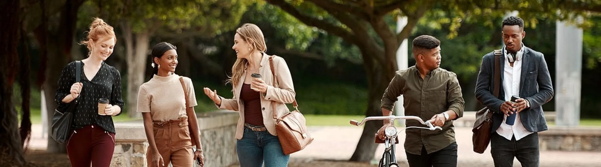 Five friends enjoying a sunny walk in a park, engaging in lively conversation, with one person walking a bicycle.