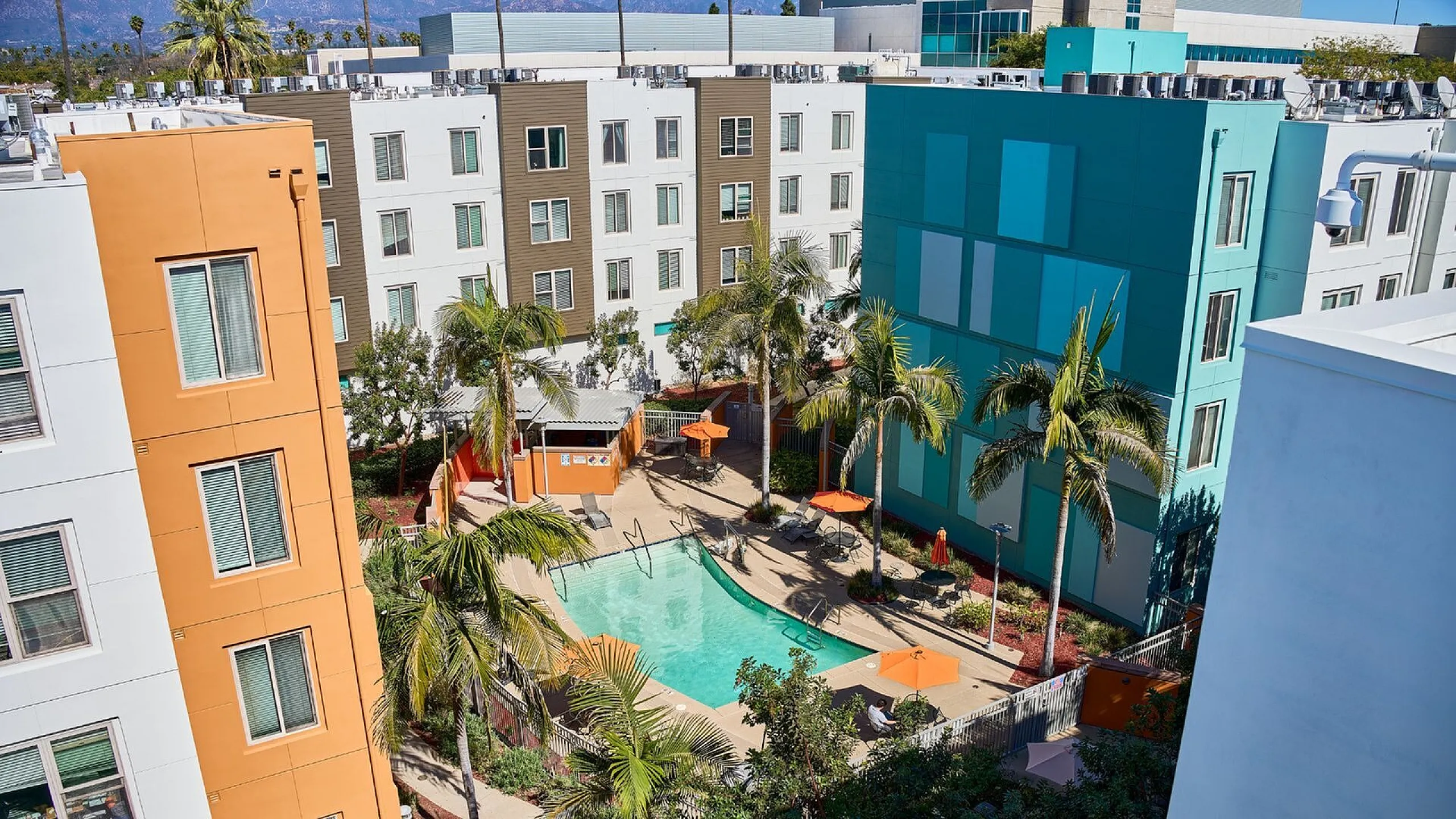 Modern apartment complex with vibrant colors overlooking a sunlit courtyard with palm trees and a sparkling swimming pool.