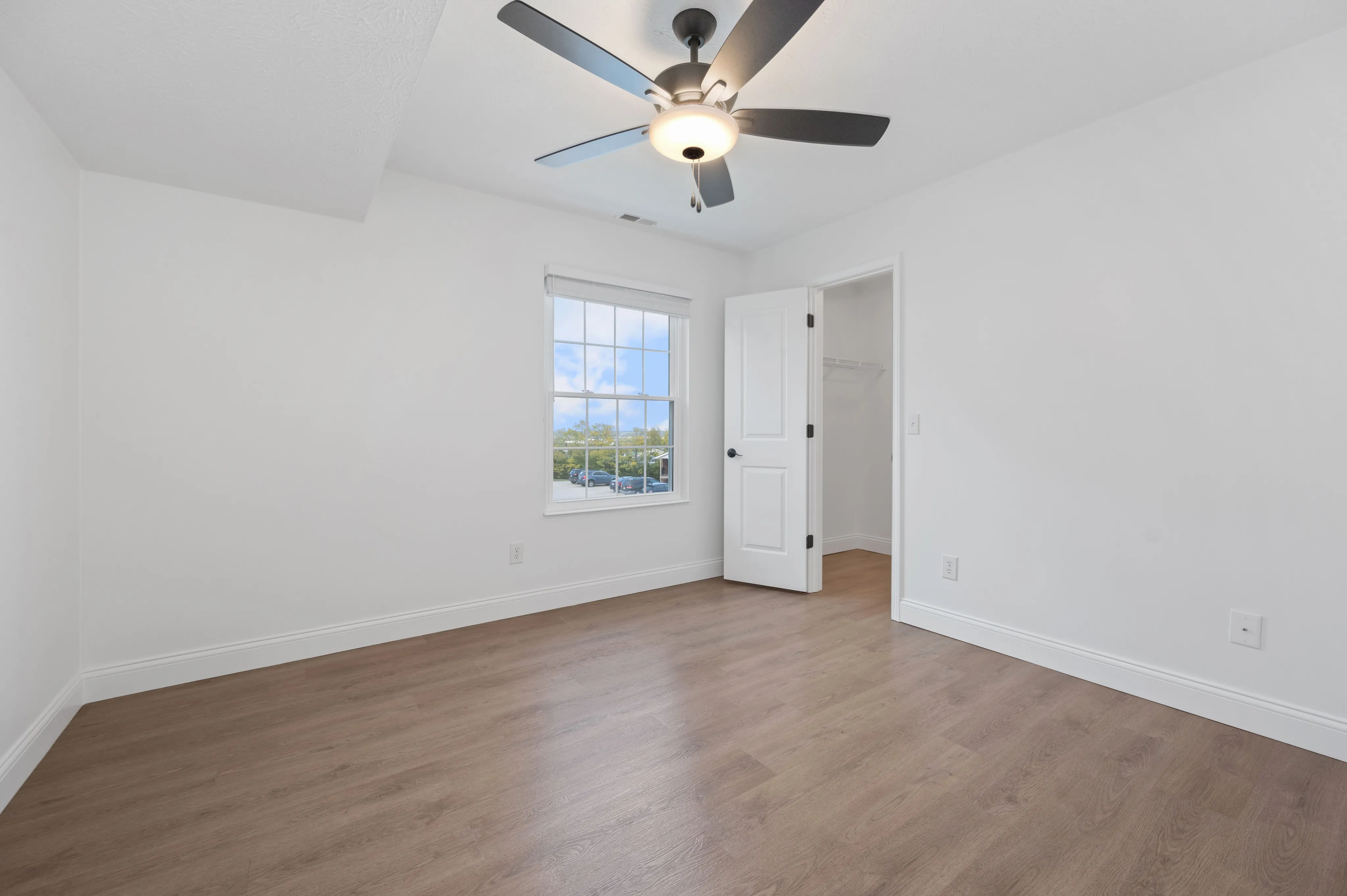 Spacious bedroom with wooden flooring, large window for natural light, and ceiling fan.