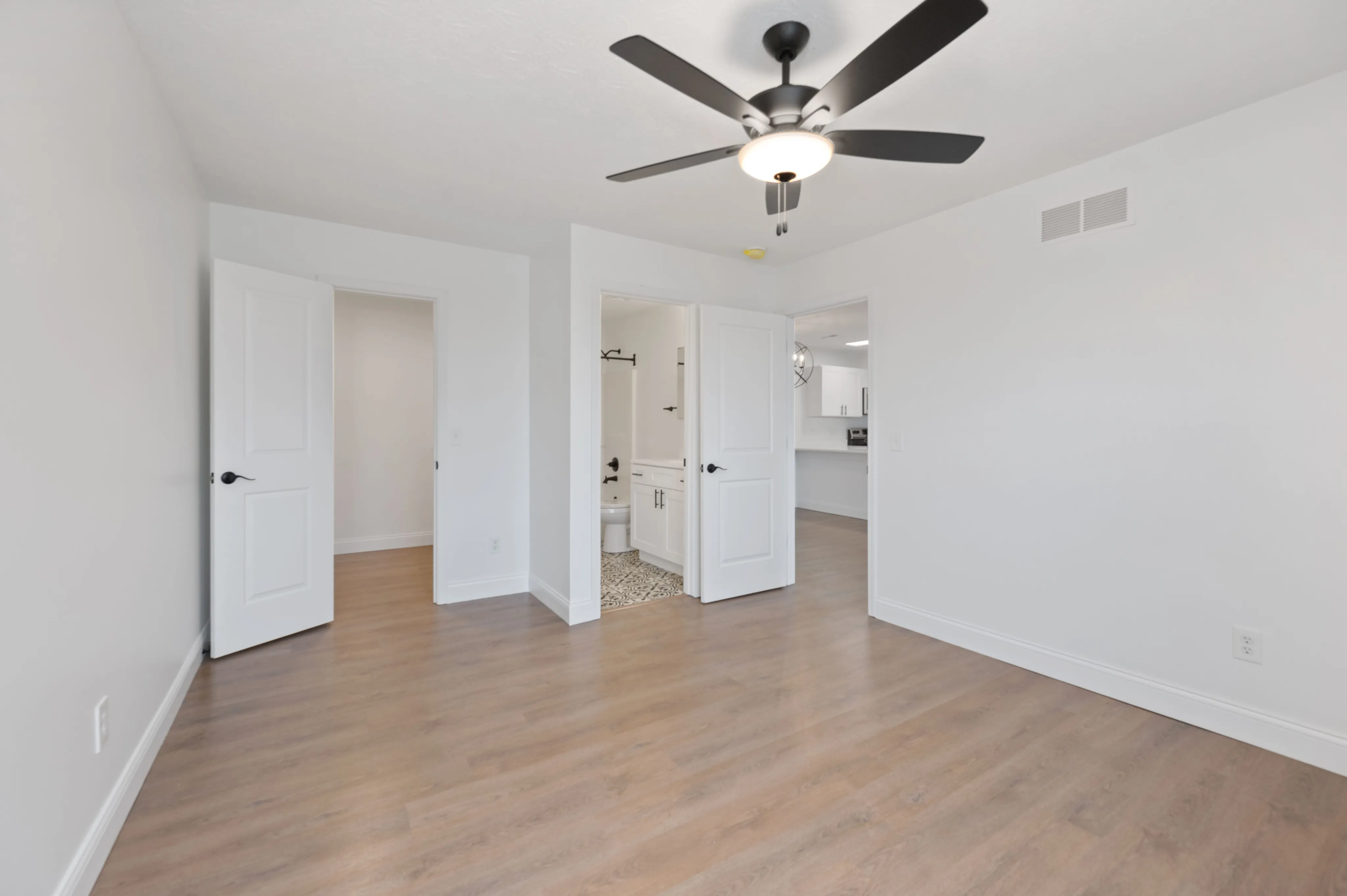 Spacious bedroom with wooden flooring, ceiling fan, and open doors leading to a bathroom and kitchen.