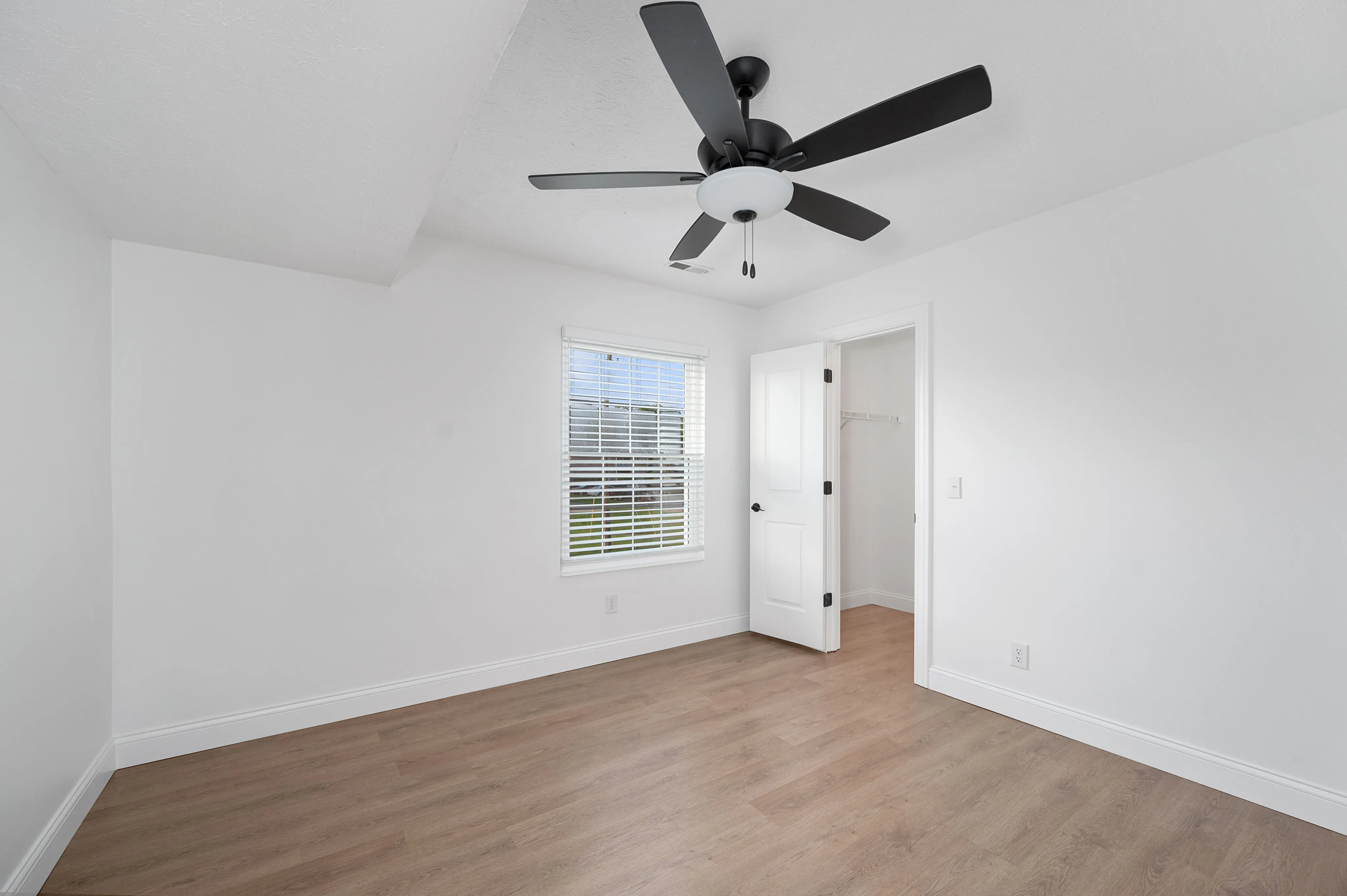 A bright, airy bedroom with a modern ceiling fan and hardwood flooring, featuring a large window and an open closet.