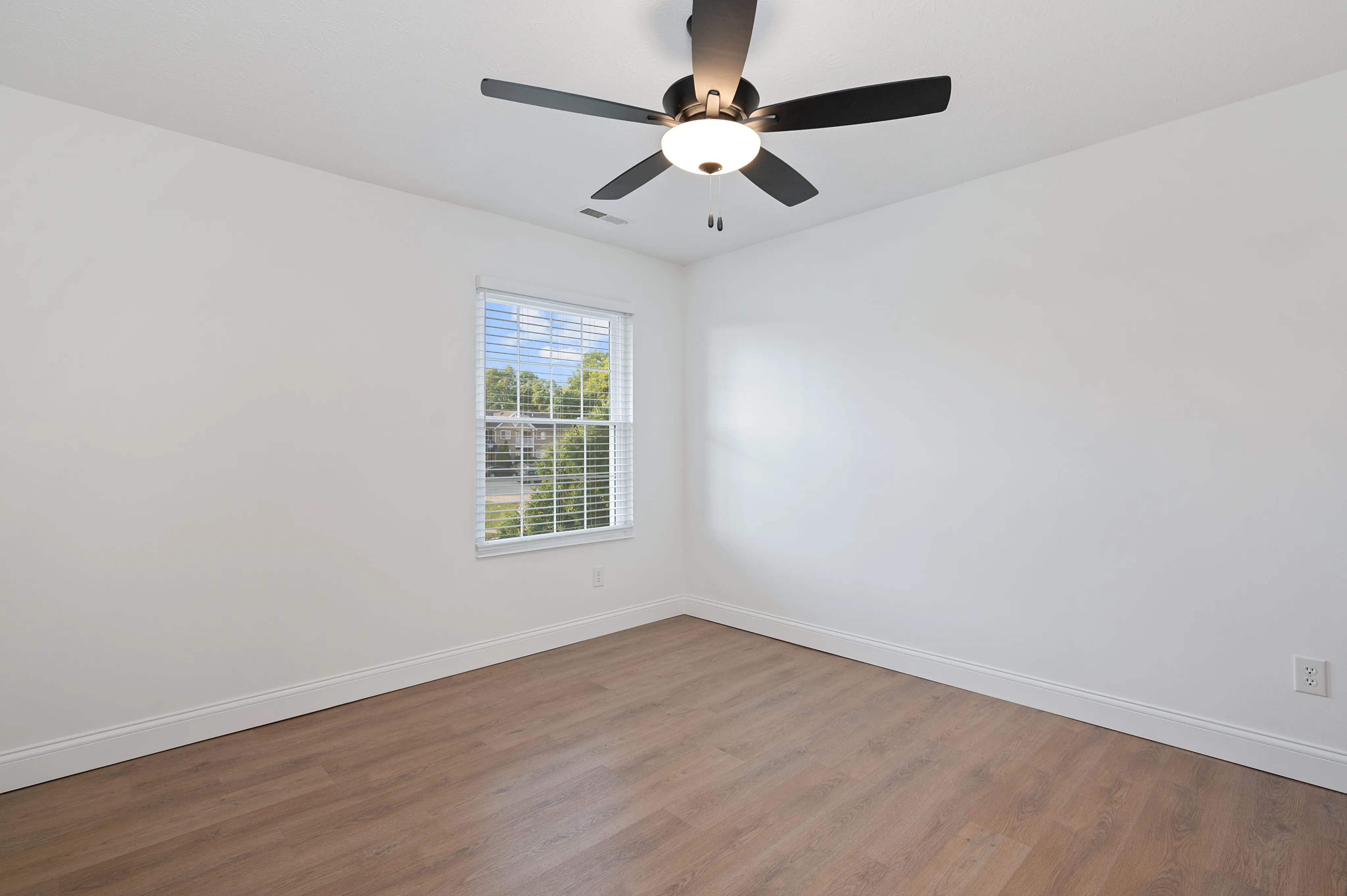 Spacious, empty bedroom with wooden flooring and a modern ceiling fan, illuminated by natural light from a large window.