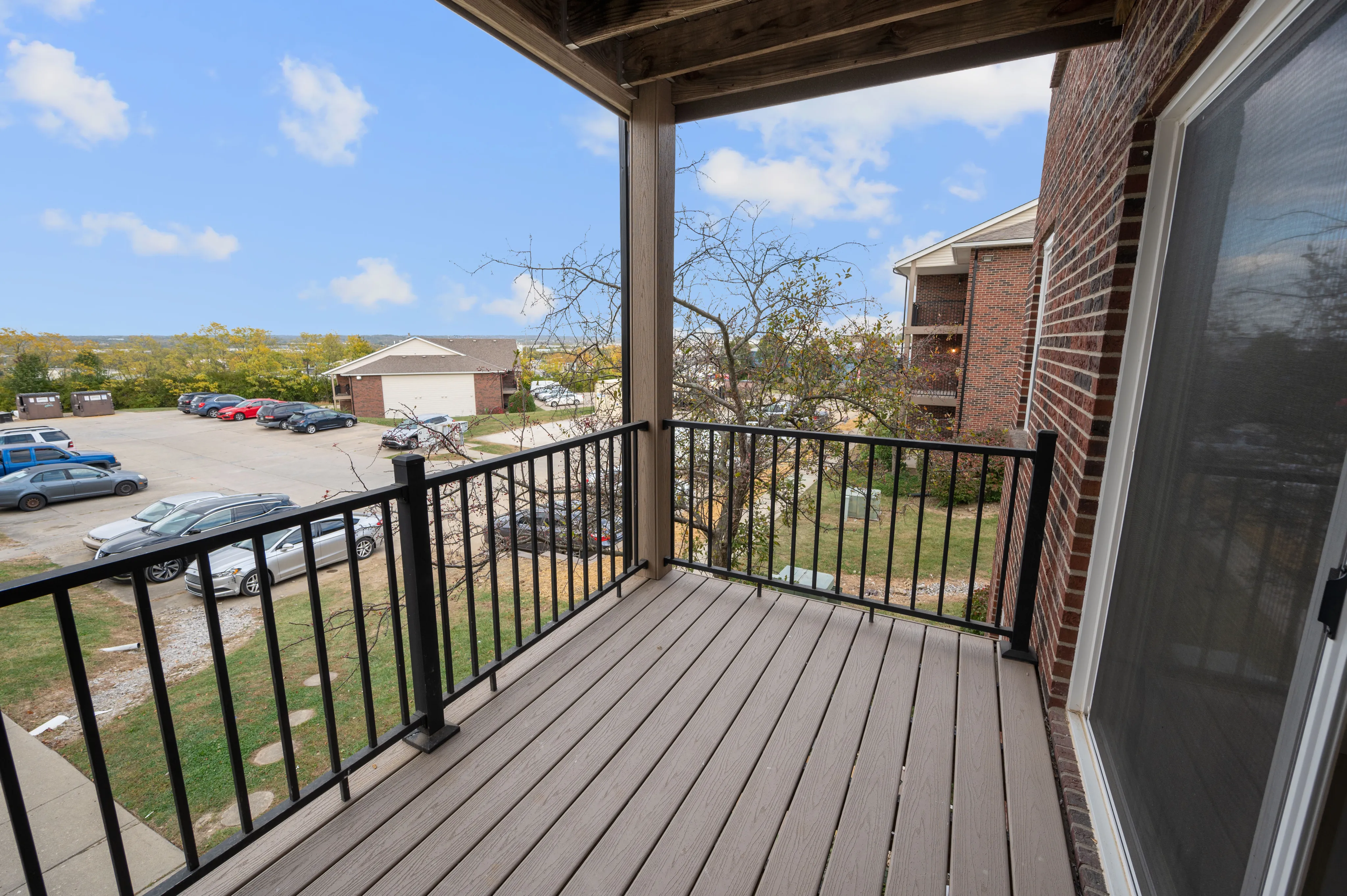 Inviting balcony with a wooden deck overlooking a parking area and nearby buildings, set against a clear blue sky.