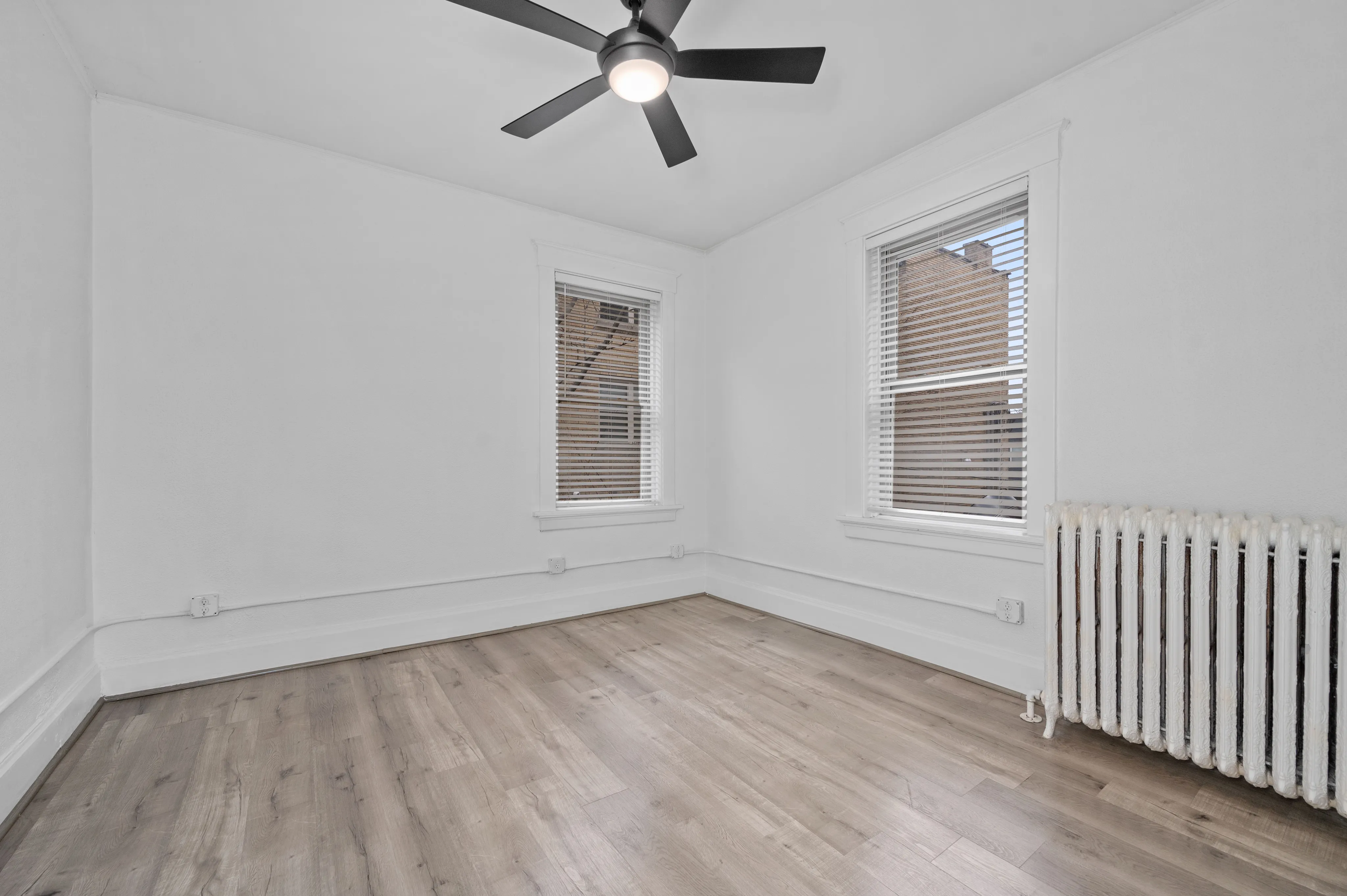 Bright and airy empty room featuring sleek modern ceiling fan, large windows with blinds, and elegant wood flooring.