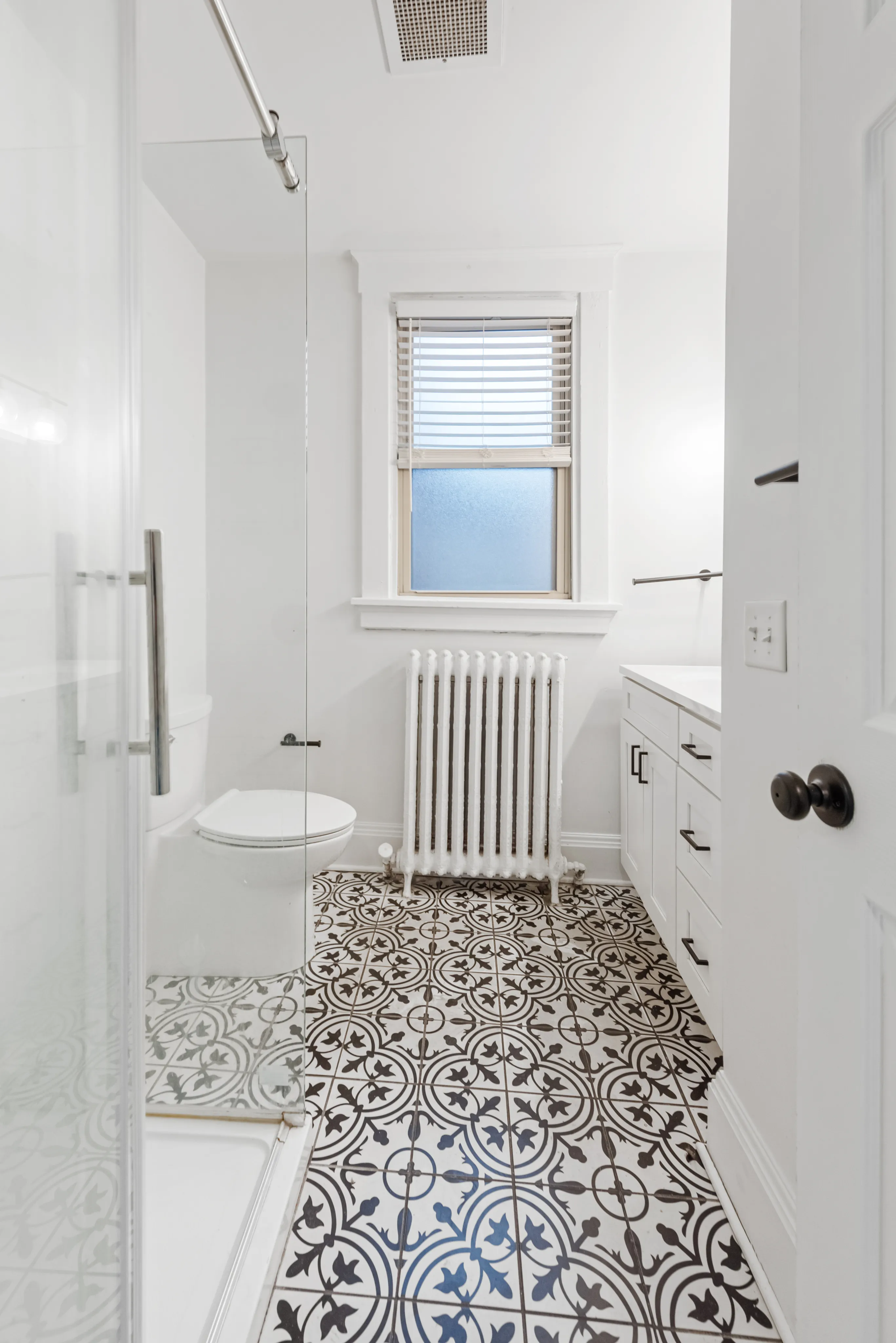 Charming bathroom with elegant patterned tile flooring, sleek glass shower, and classic radiator under a bright window.