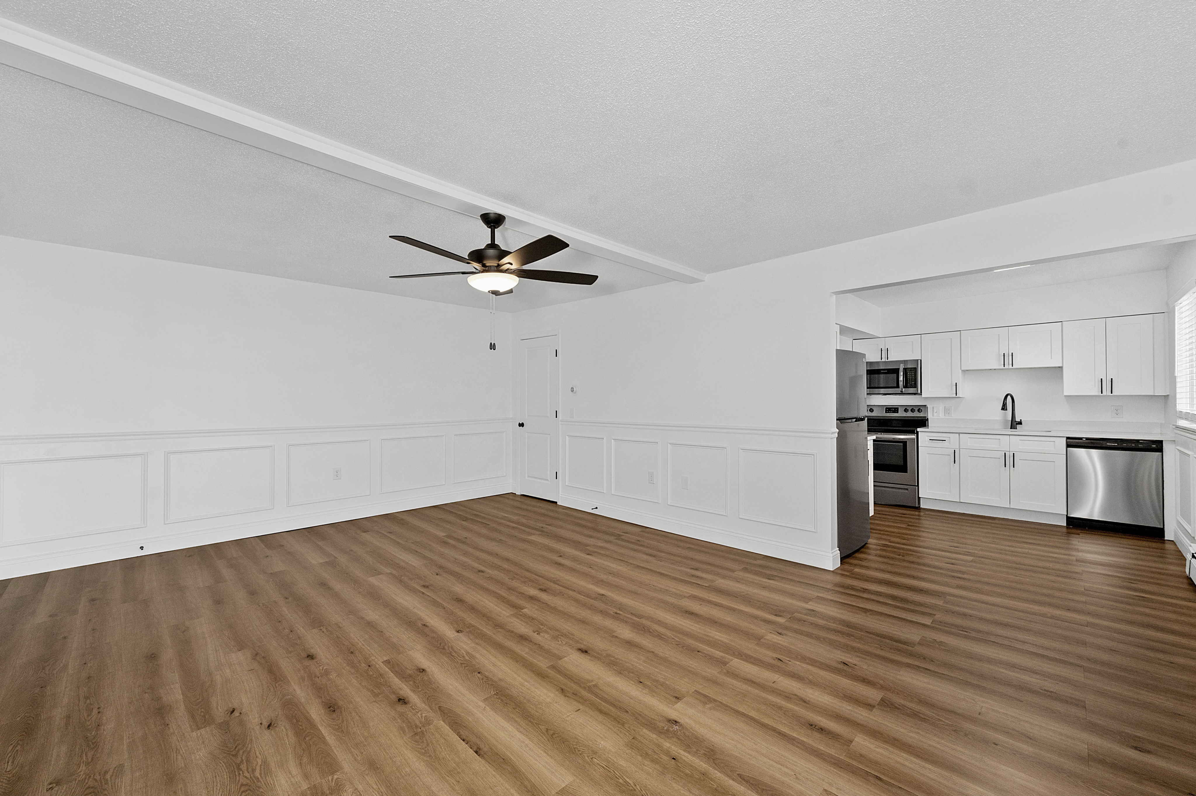 Modern open-plan living room with elegant flooring, sleek ceiling fan, and contemporary kitchen featuring stainless steel appliances and white cabinetry.