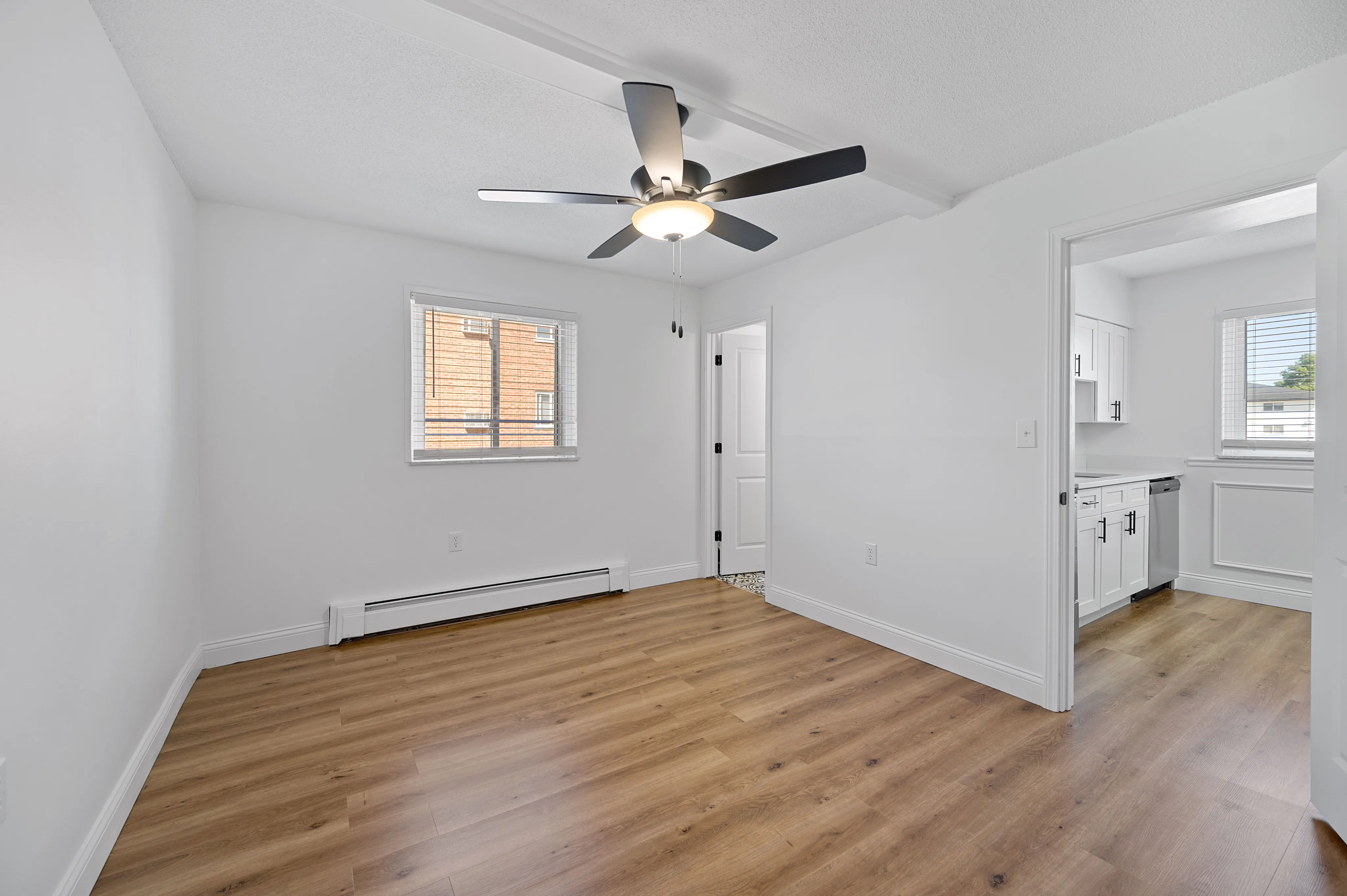 Bright, spacious room with modern ceiling fan, sleek wooden flooring, and access to a cozy kitchen in a stylish apartment.