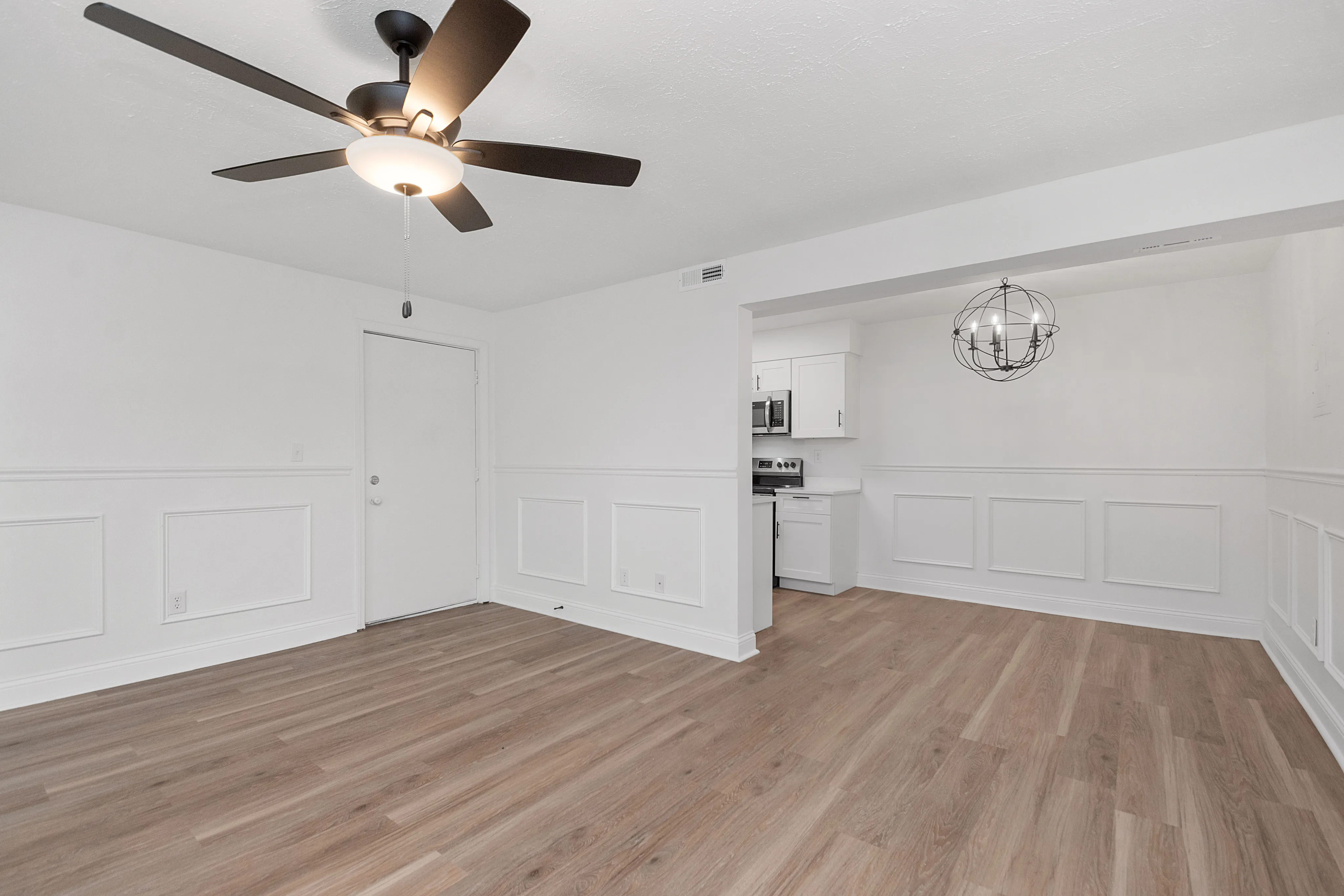 Bright and modern open-plan living room with elegant light fixtures, hardwood floors, and a peek into a sleek kitchen.