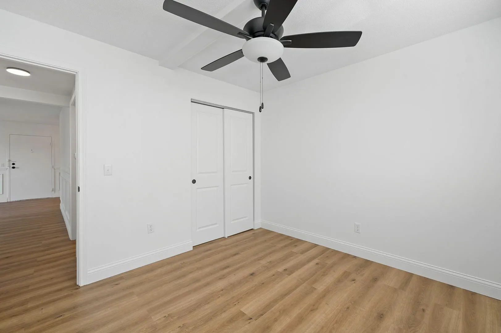 Bright and inviting room with sleek wooden flooring, modern ceiling fan, and pristine white walls, perfect for a stylish living space.