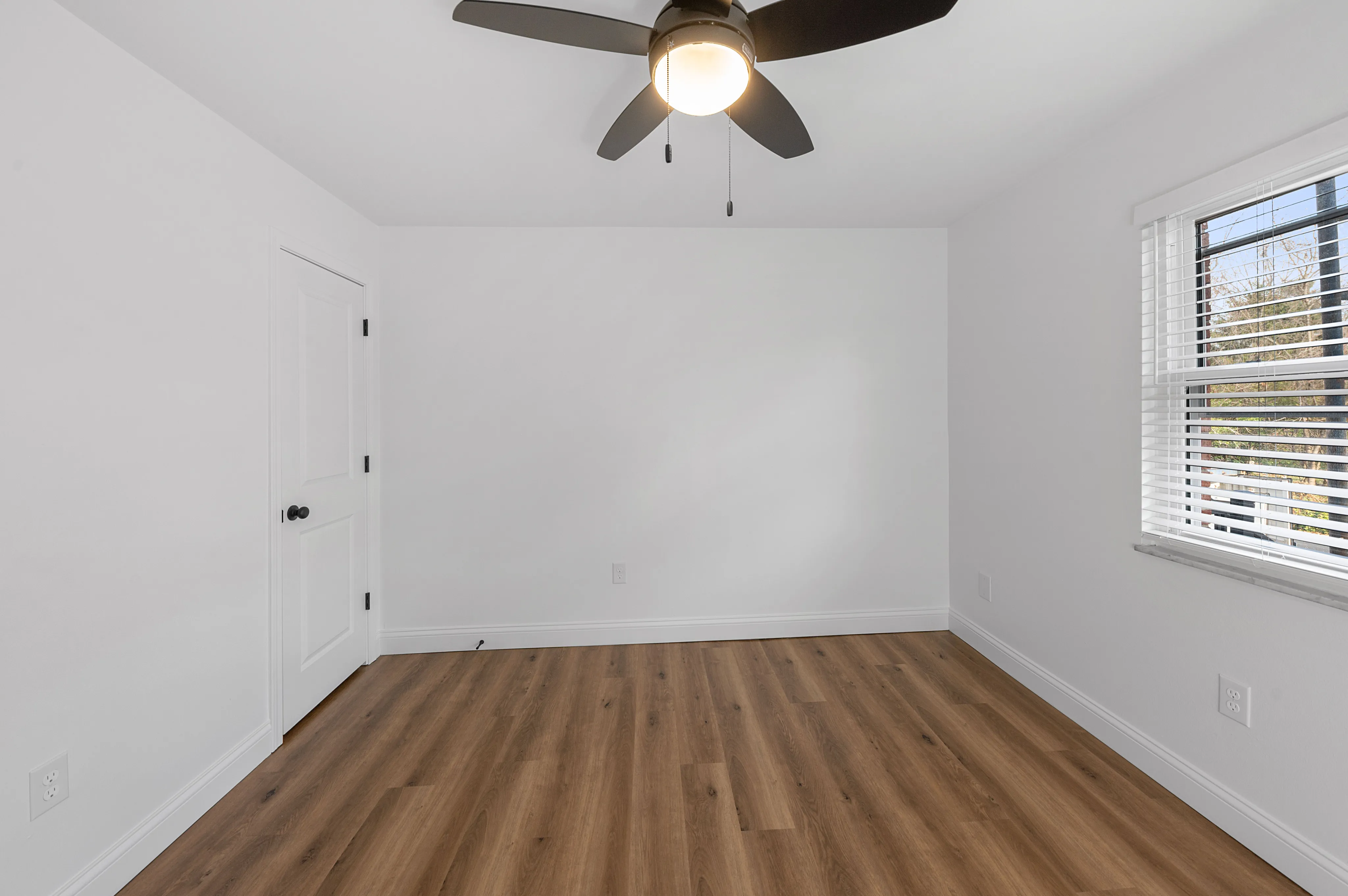 Bright, empty bedroom with a modern ceiling fan, chic hardwood floors, and large window flooding the room with natural light.