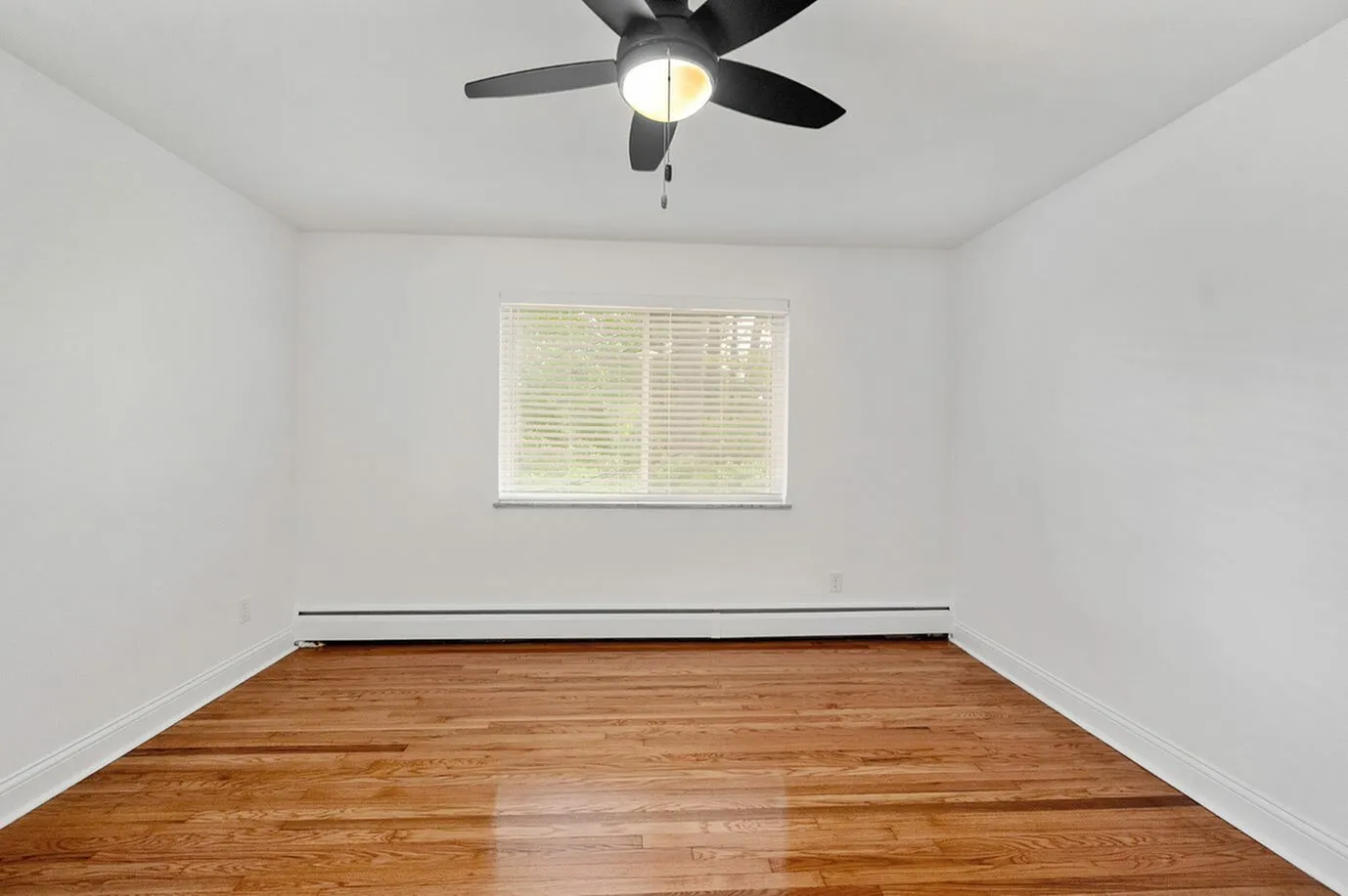 Bright and airy empty room with polished hardwood floors, modern ceiling fan, and large window offering natural light, perfect for a cozy bedroom or home office.