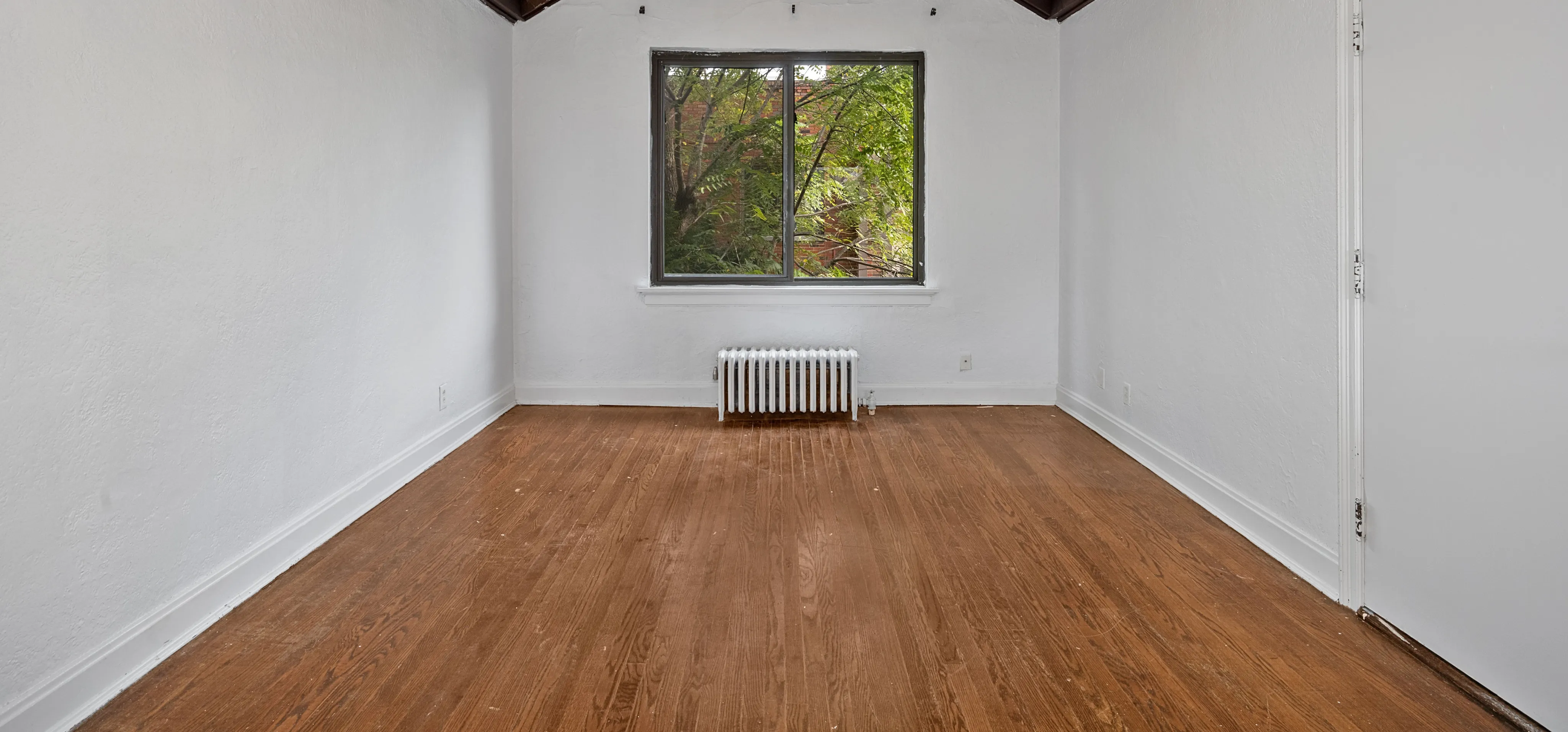 Bright room with hardwood floors, large window, and fresh white walls offering a serene view of greenery.
