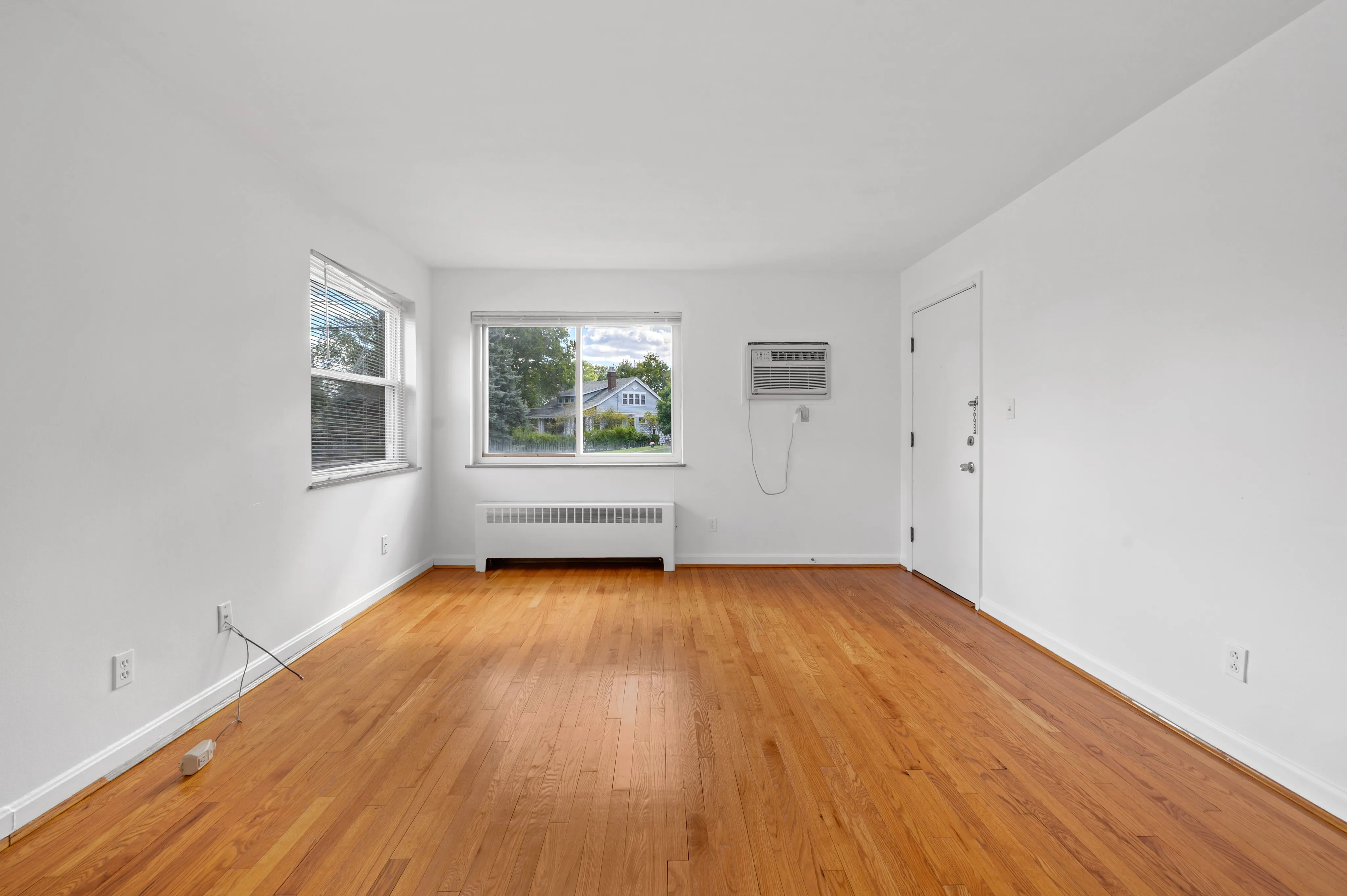 Bright living room with large window, polished hardwood floors, and modern wall unit air conditioner.