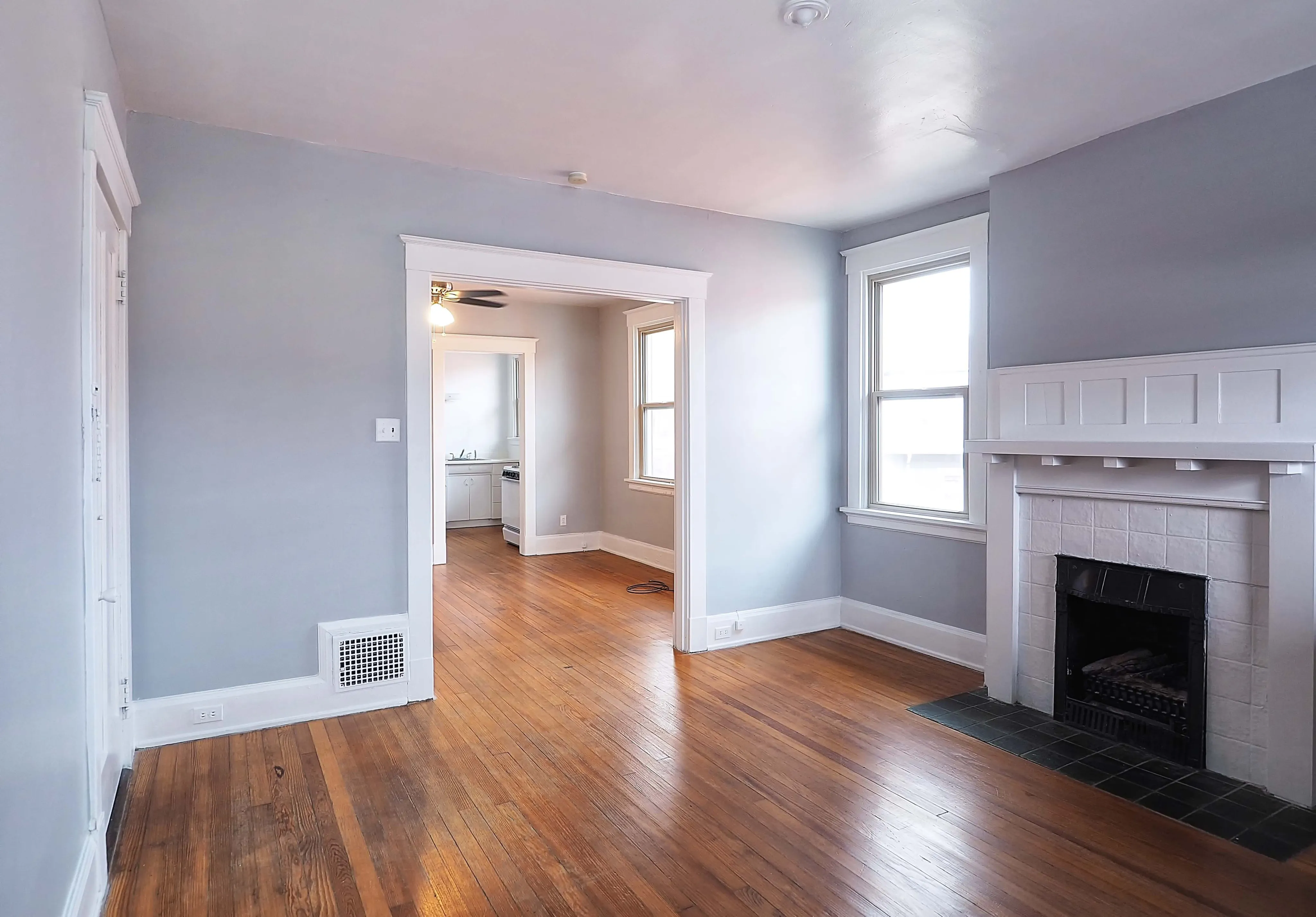 Bright living room with hardwood floors, large windows, and a classic fireplace, leading into a cozy kitchen area.