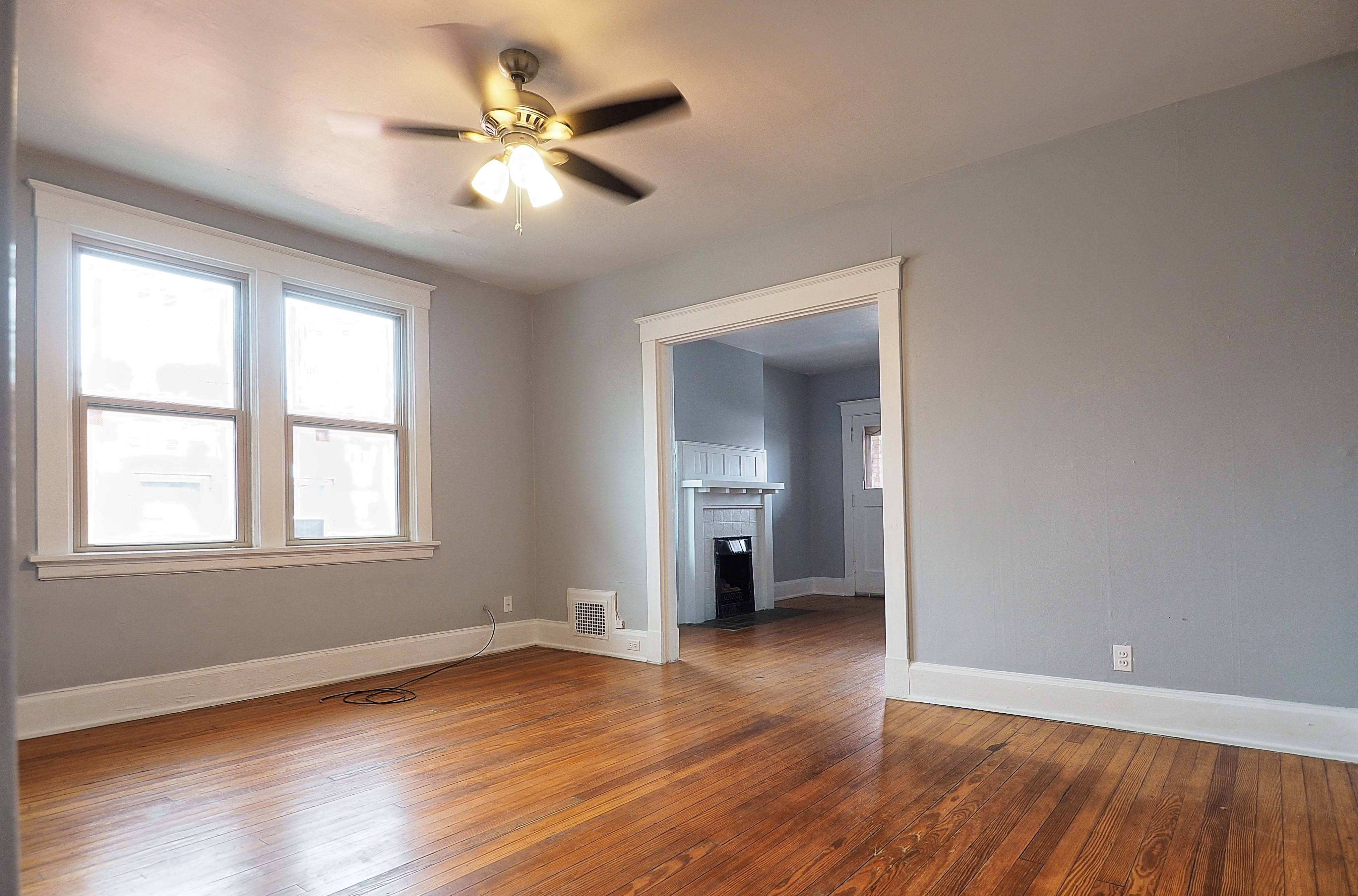 Charming living space with warm hardwood floors, a ceiling fan, and bright windows leading to a cozy room with a fireplace.