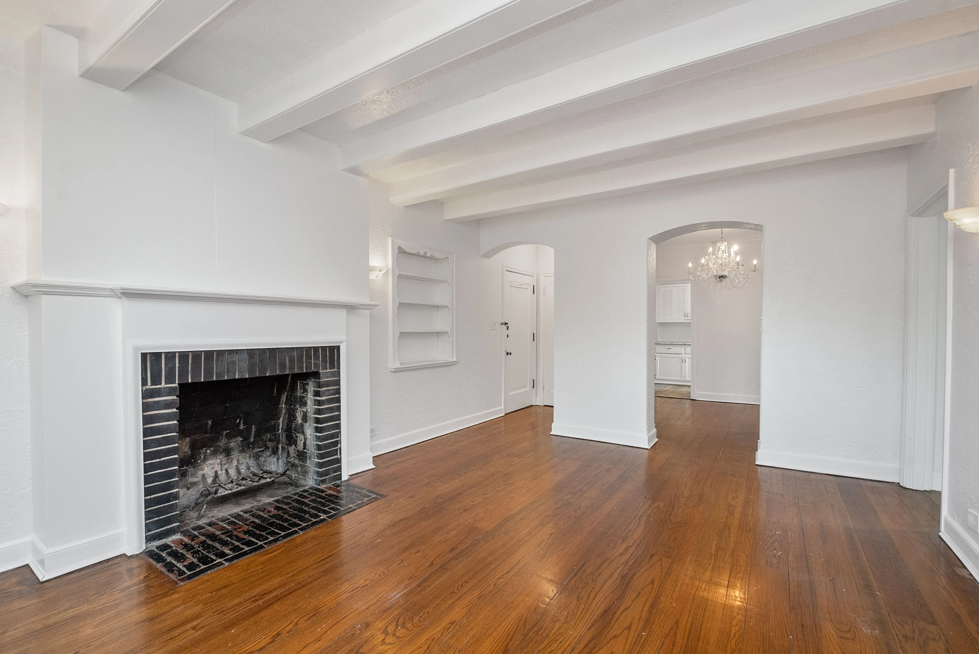 Elegant living room with exposed beams, polished hardwood floors, and a charming fireplace, leading to an inviting dining area with a stylish chandelier.