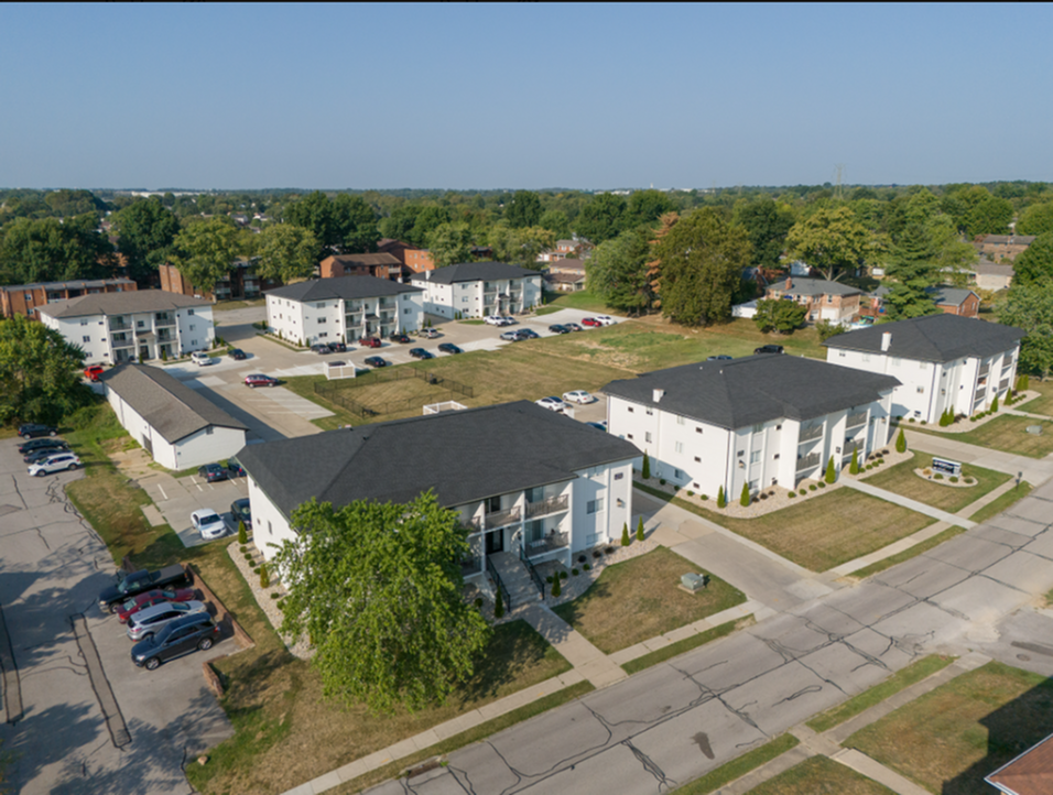 Aerial view of a modern apartment complex surrounded by greenery, showcasing multiple white buildings with spacious parking and landscaped pathways.