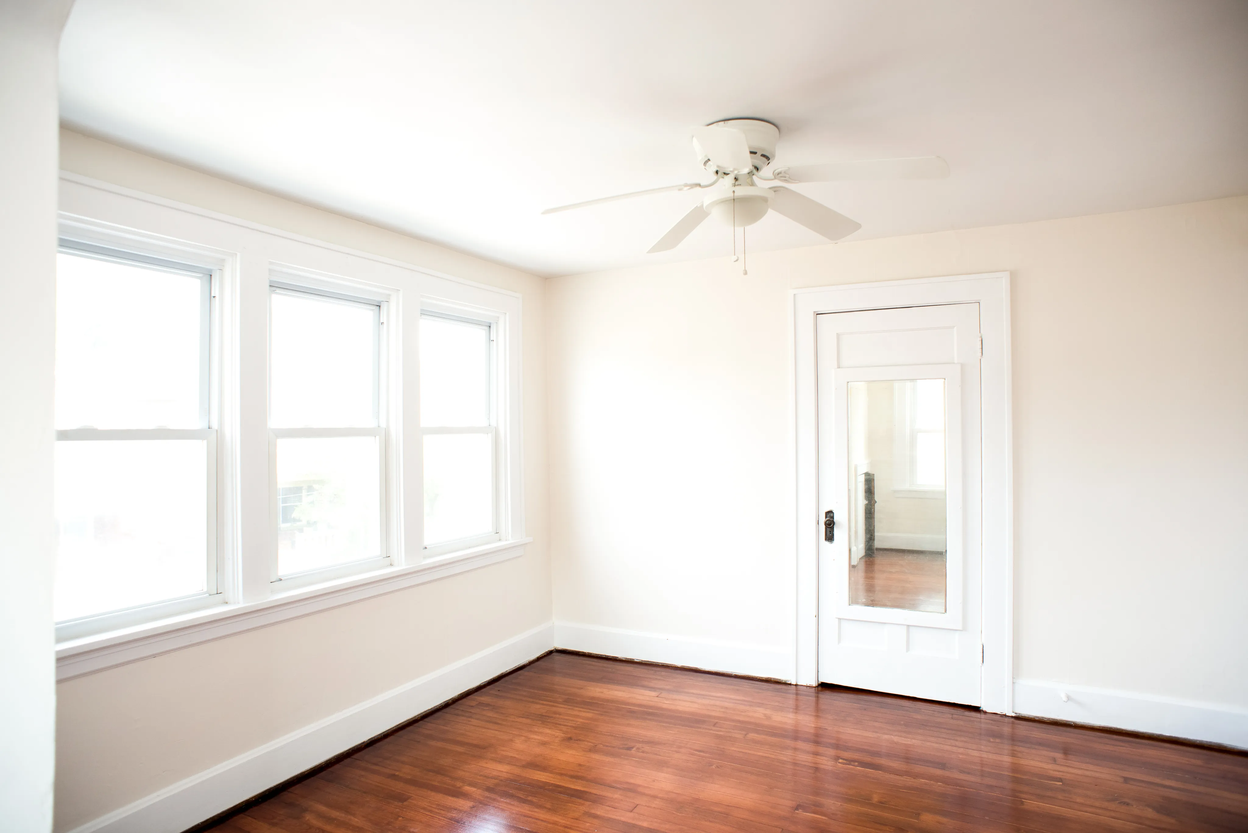 Bright room with elegant hardwood floors and large windows, featuring a sleek ceiling fan and a stylish door with mirror.