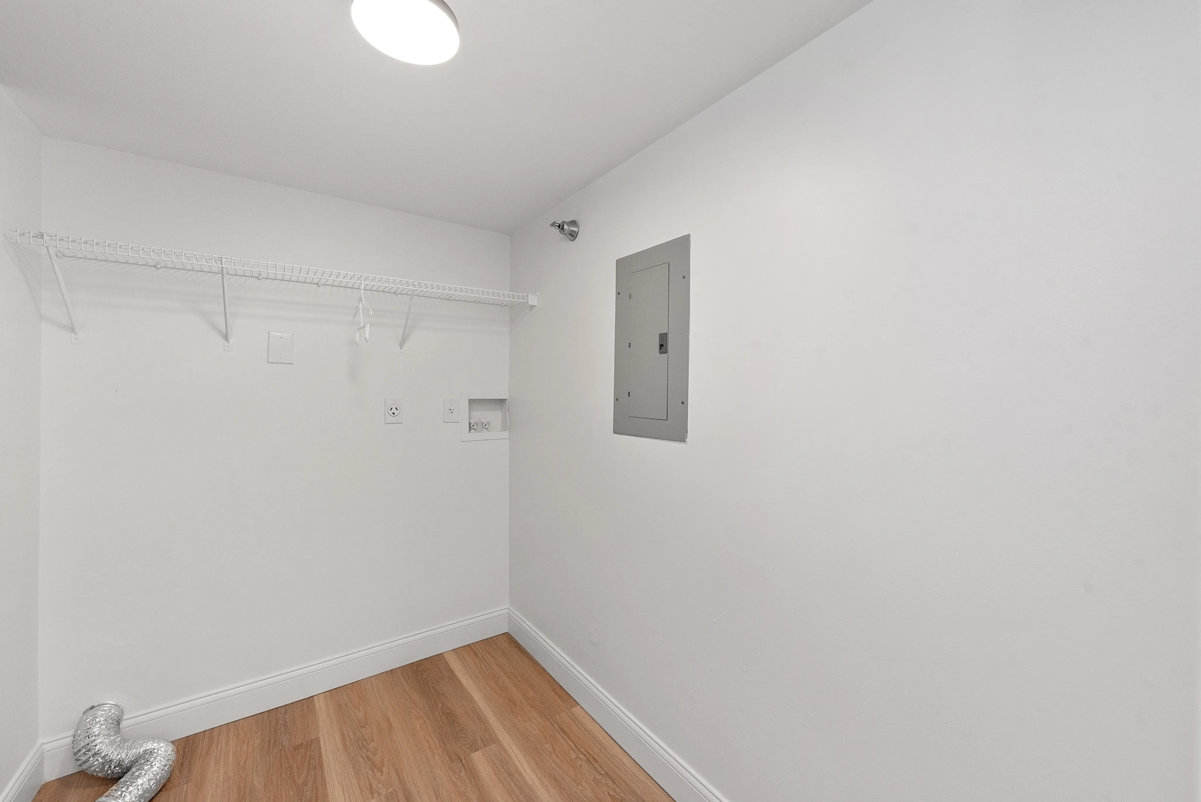 Bright laundry room with wood flooring, wall shelving, and utility connections.
