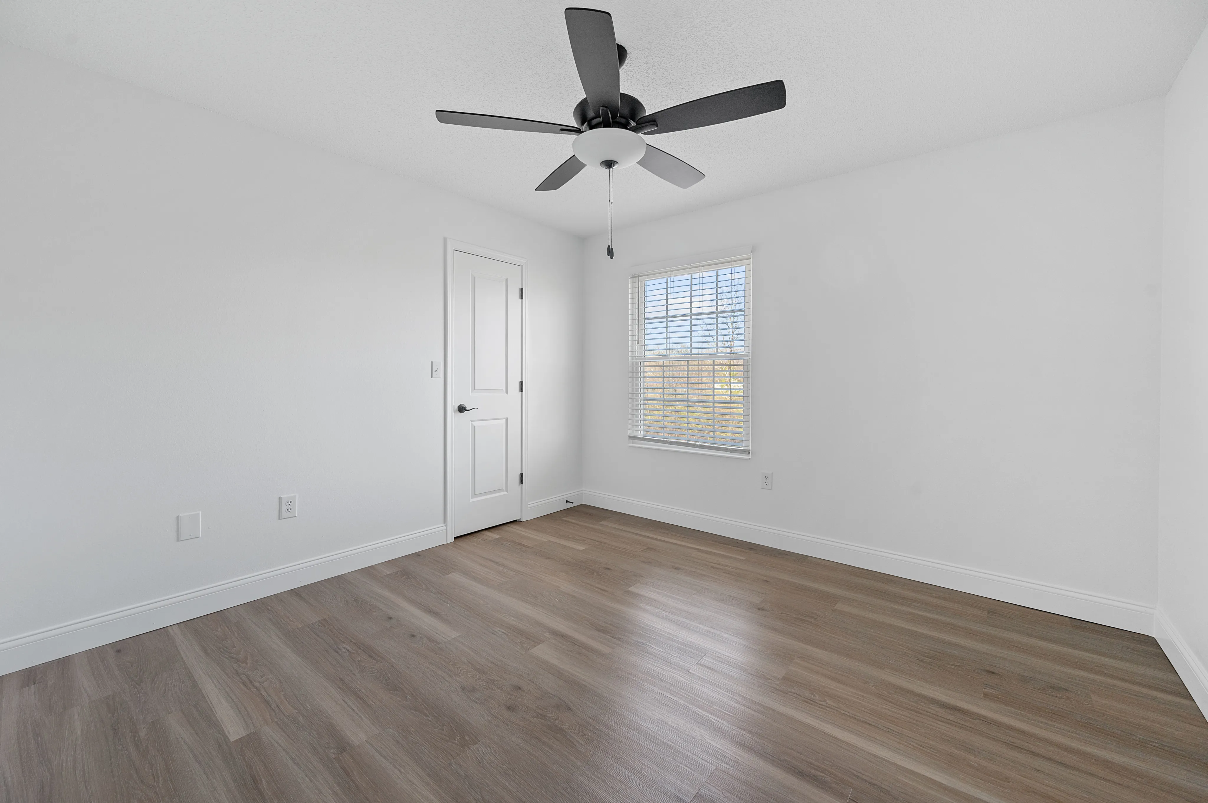 Bright, sunlit bedroom featuring sleek hardwood floors and a modern ceiling fan, offering a spacious and airy atmosphere.