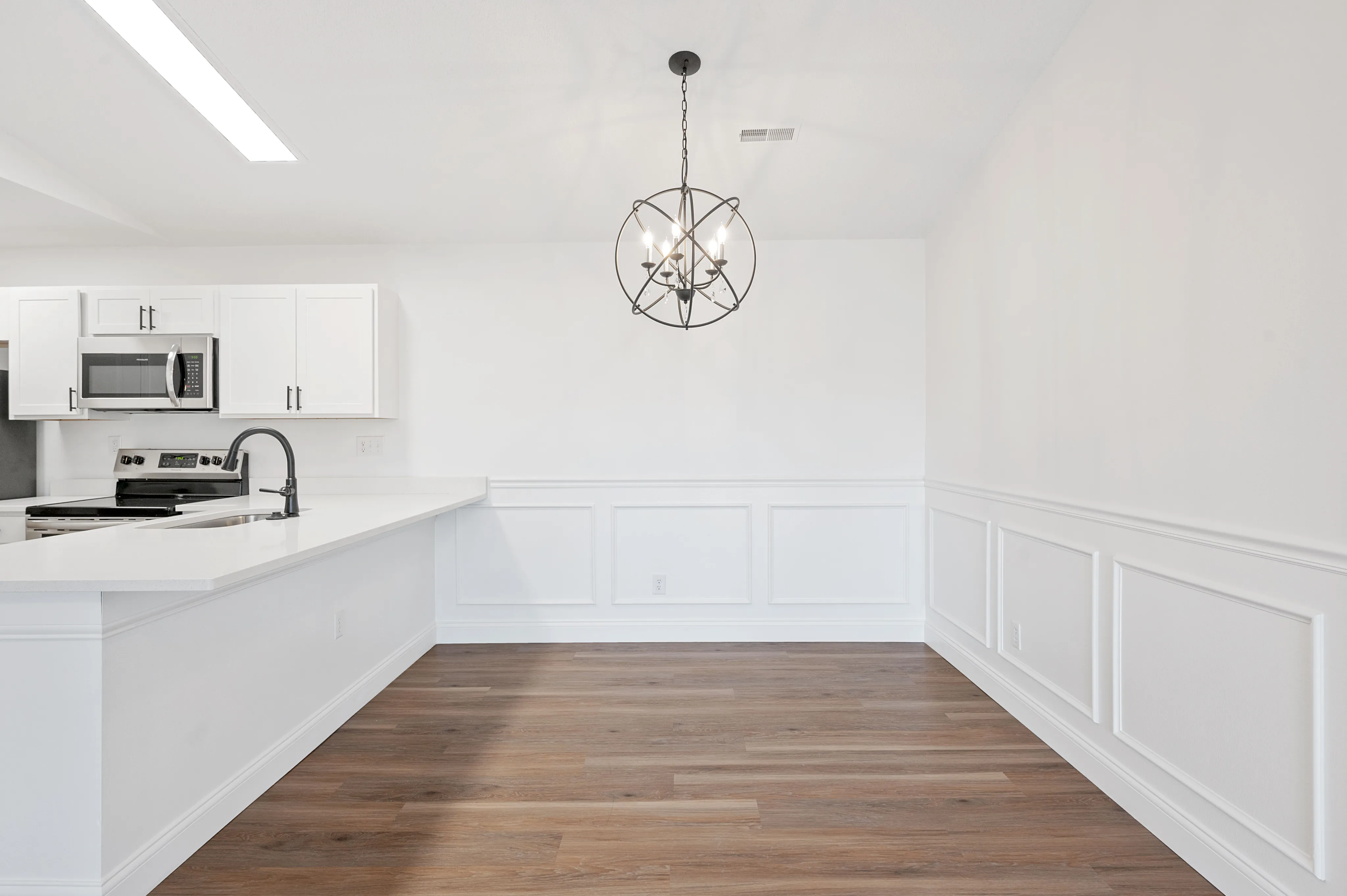 Elegant kitchen space with sleek white cabinetry, modern appliances, and a chic hanging light fixture over stylish wood flooring.