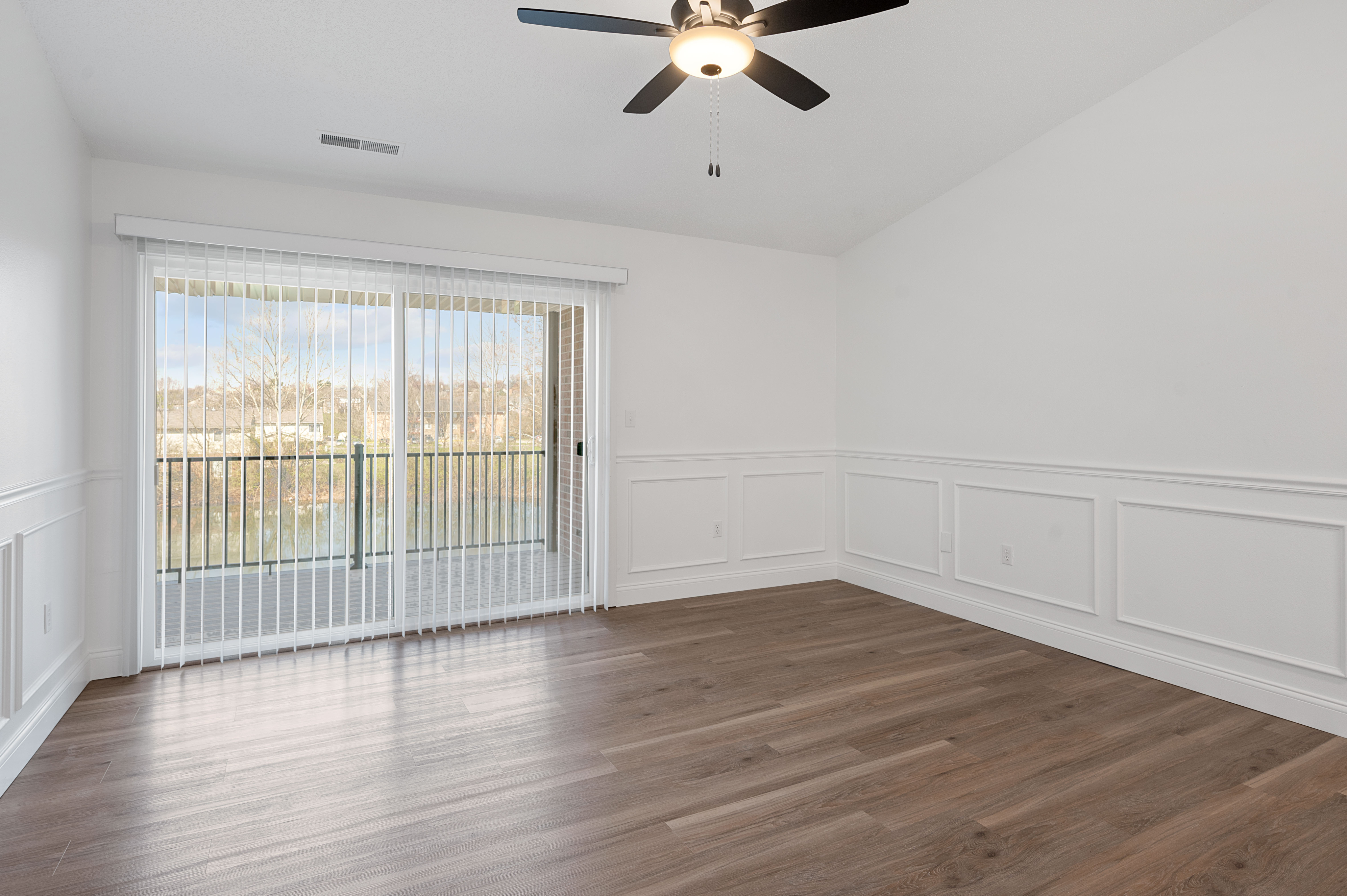 Bright, spacious room with elegant wood flooring, ceiling fan, and floor-to-ceiling sliding glass doors leading to a serene balcony view.