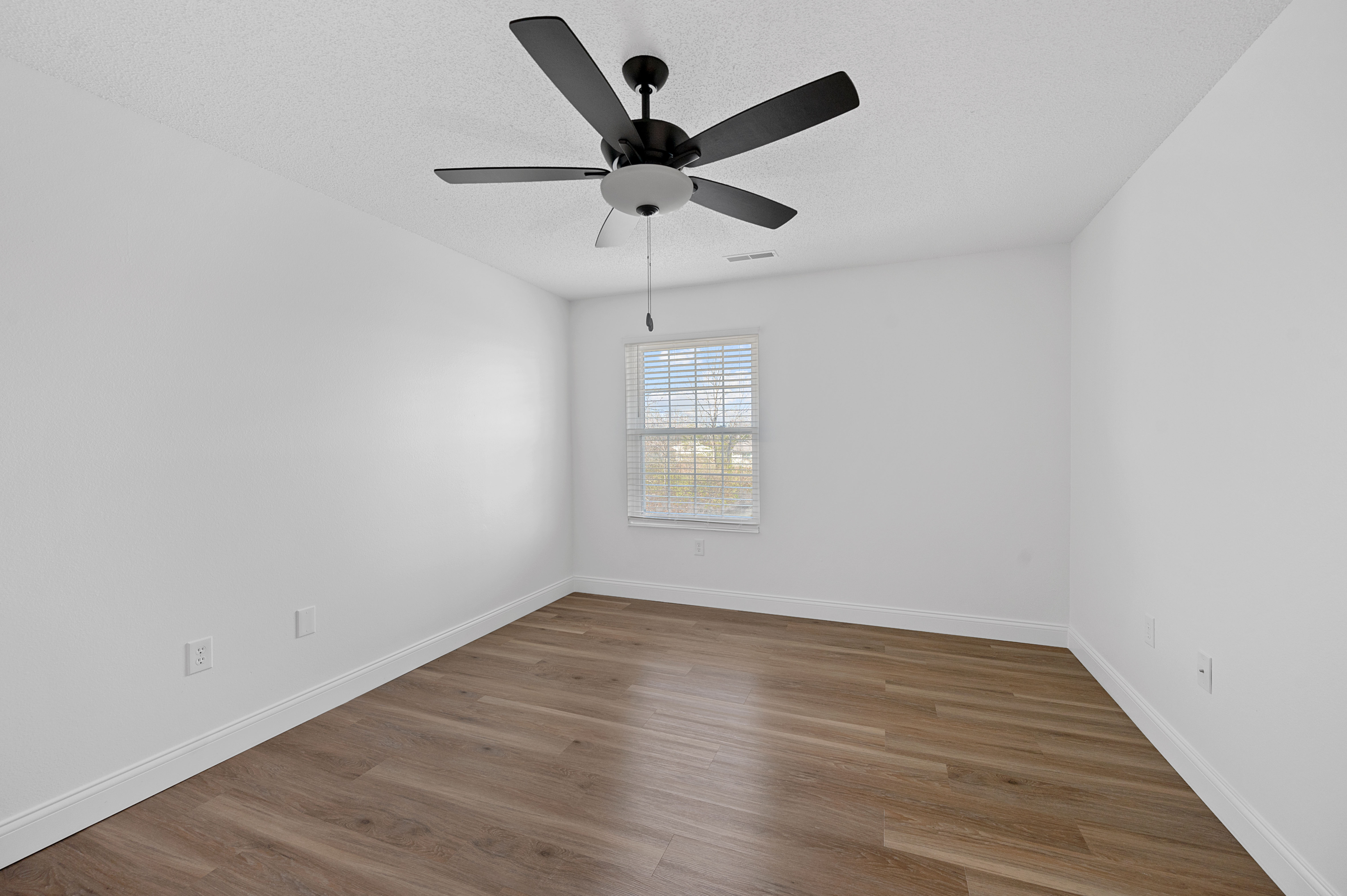 Sunny, spacious bedroom with elegant wooden flooring, modern ceiling fan, and large window inviting natural light.
