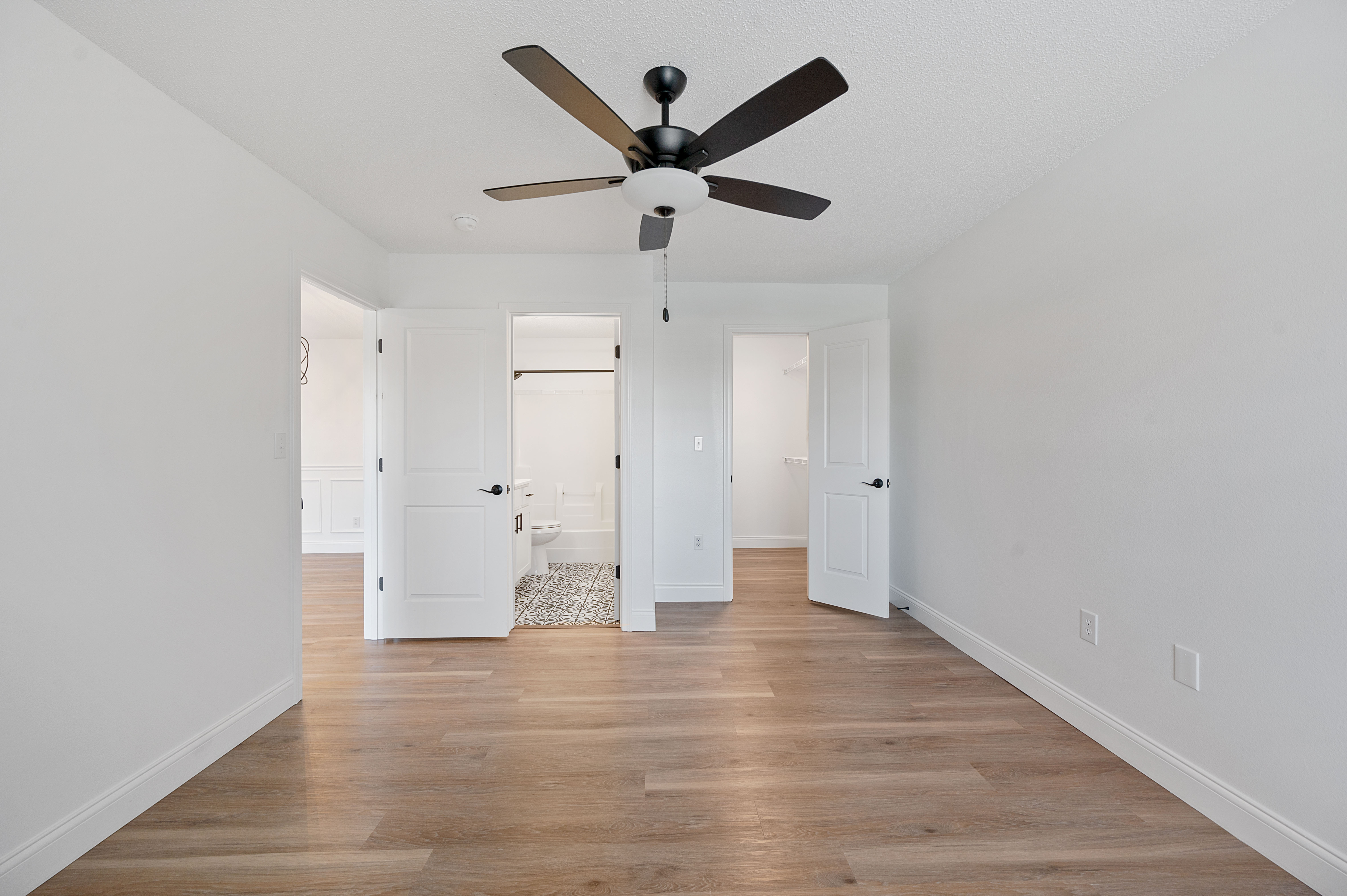 Bright bedroom with elegant wooden flooring, ceiling fan, and dual doors leading to a modern bathroom and spacious closet.