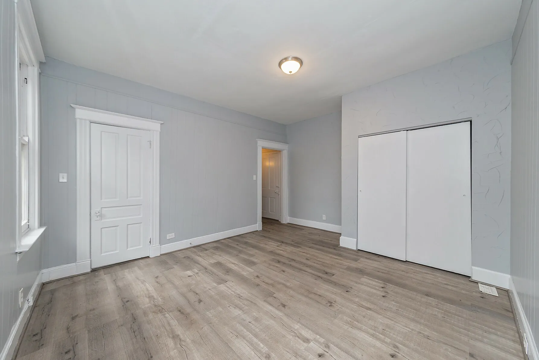Bright and spacious room with elegant wood flooring, soft gray walls, and ample natural light from a large window.