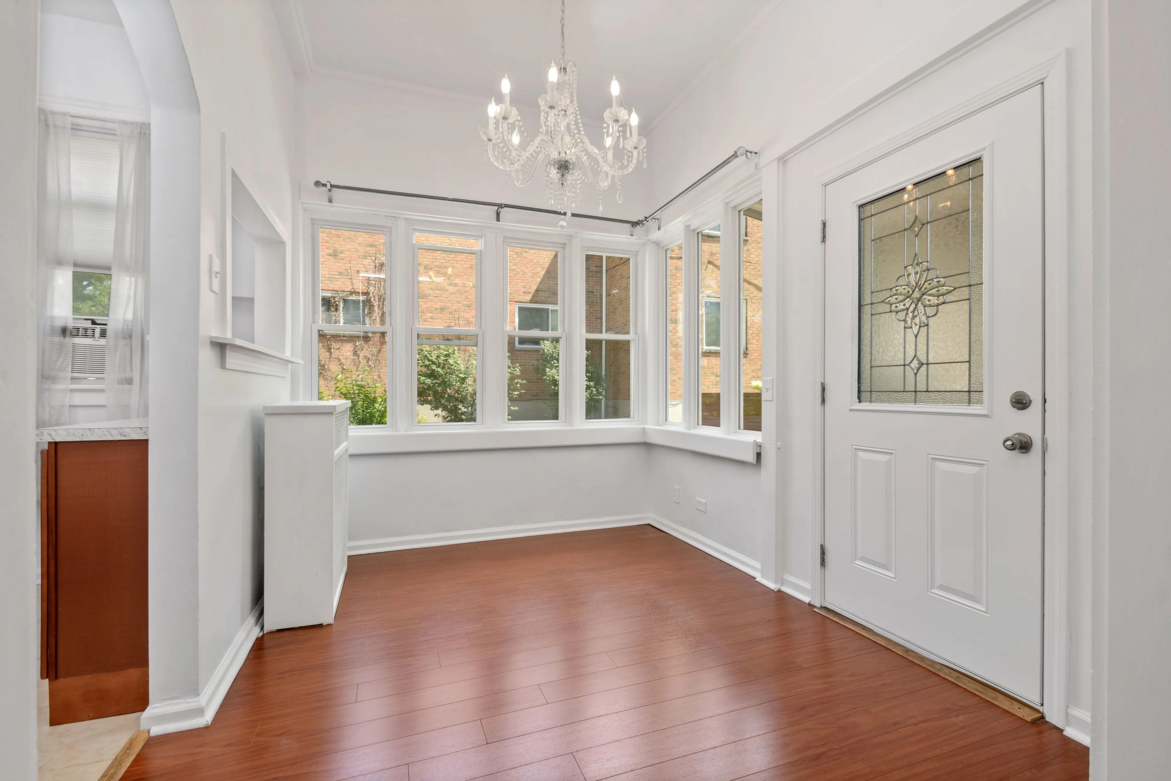 Bright and inviting corner space with elegant chandelier, large windows, and polished wood flooring, perfect for dining or relaxation.